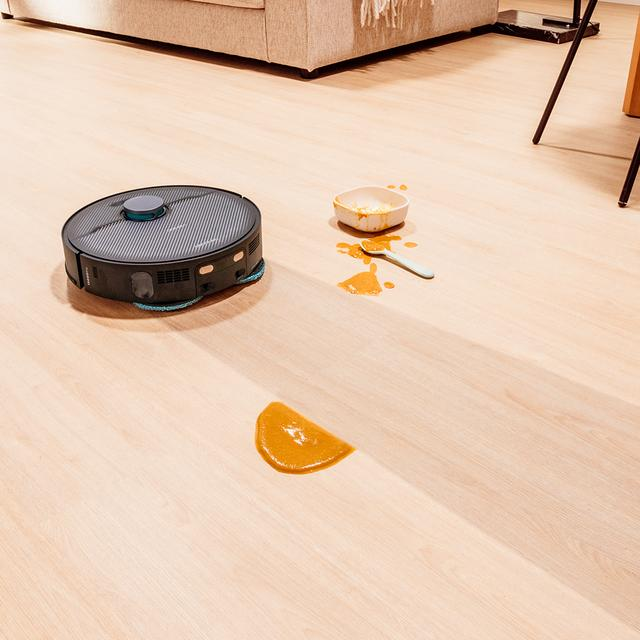 Robot süpürge, ahşap zeminde dökülmüş yiyeceklerin yanında.