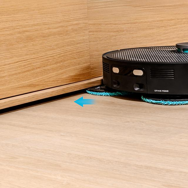 Mavi paspas pedlerine sahip siyah robot süpürge, ahşap panel duvara yakın ahşap zemini temizliyor.