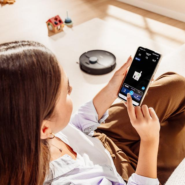 Bir kişi ahşap zeminde robot süpürgeyi akıllı telefonla kontrol ediyor.
