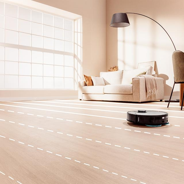 Siyah robot süpürge, koltuk ve penceresi olan bir oturma odasında açık renk ahşap zemini temizliyor.