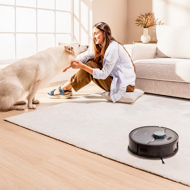 Kadın, robot süpürge ve kanepenin olduğu zeminde beyaz köpeği seviyor.