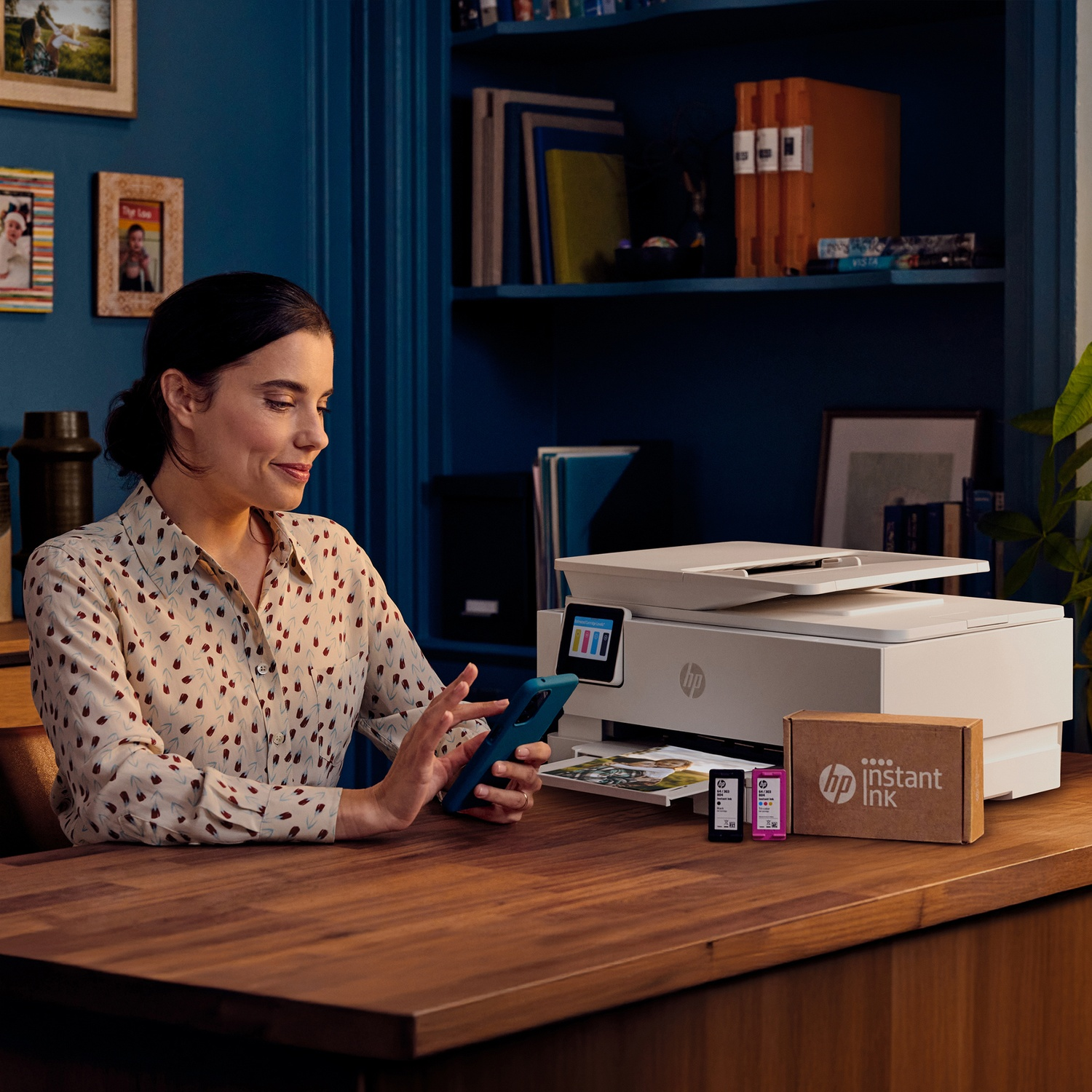 Woman uses smartphone at desk next to HP printer and Instant Ink cartridges.