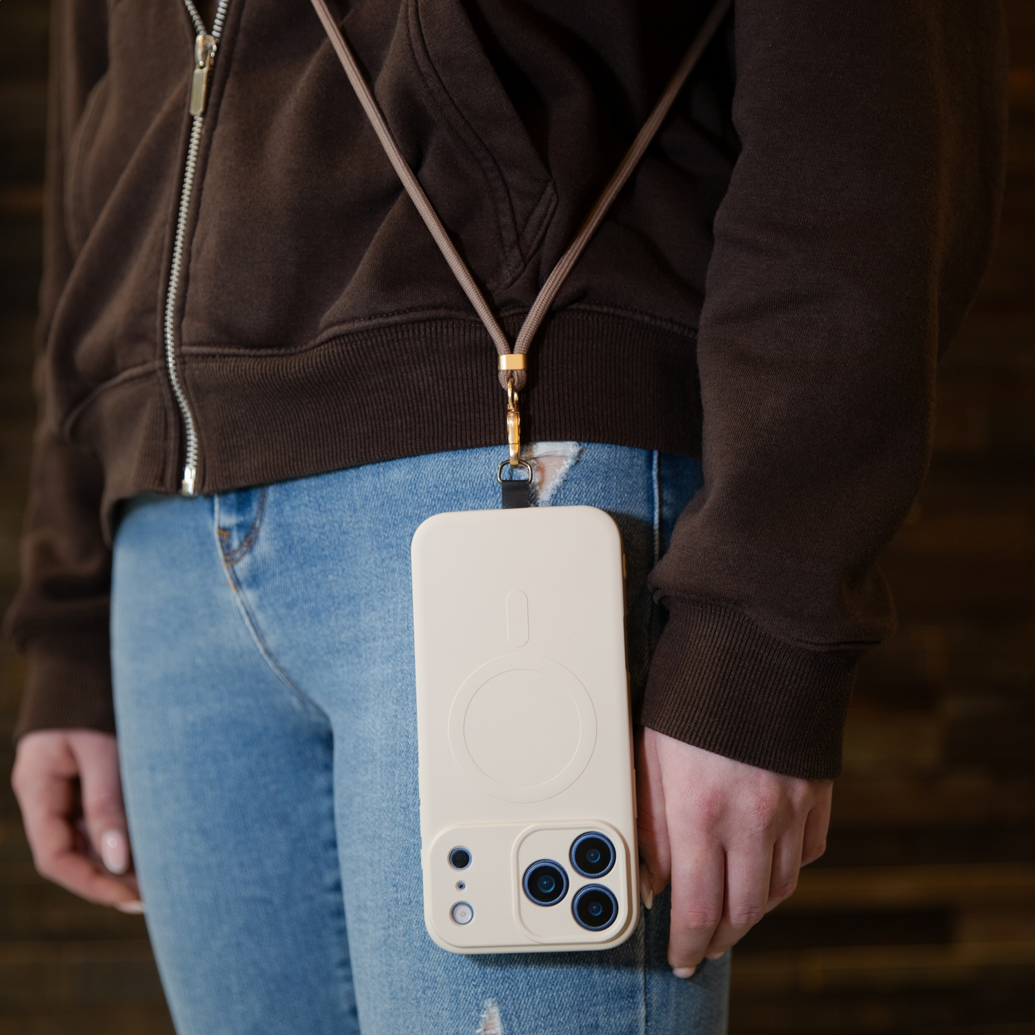 A person wears a brown hoodie, jeans, and a beige phone case with a lanyard.