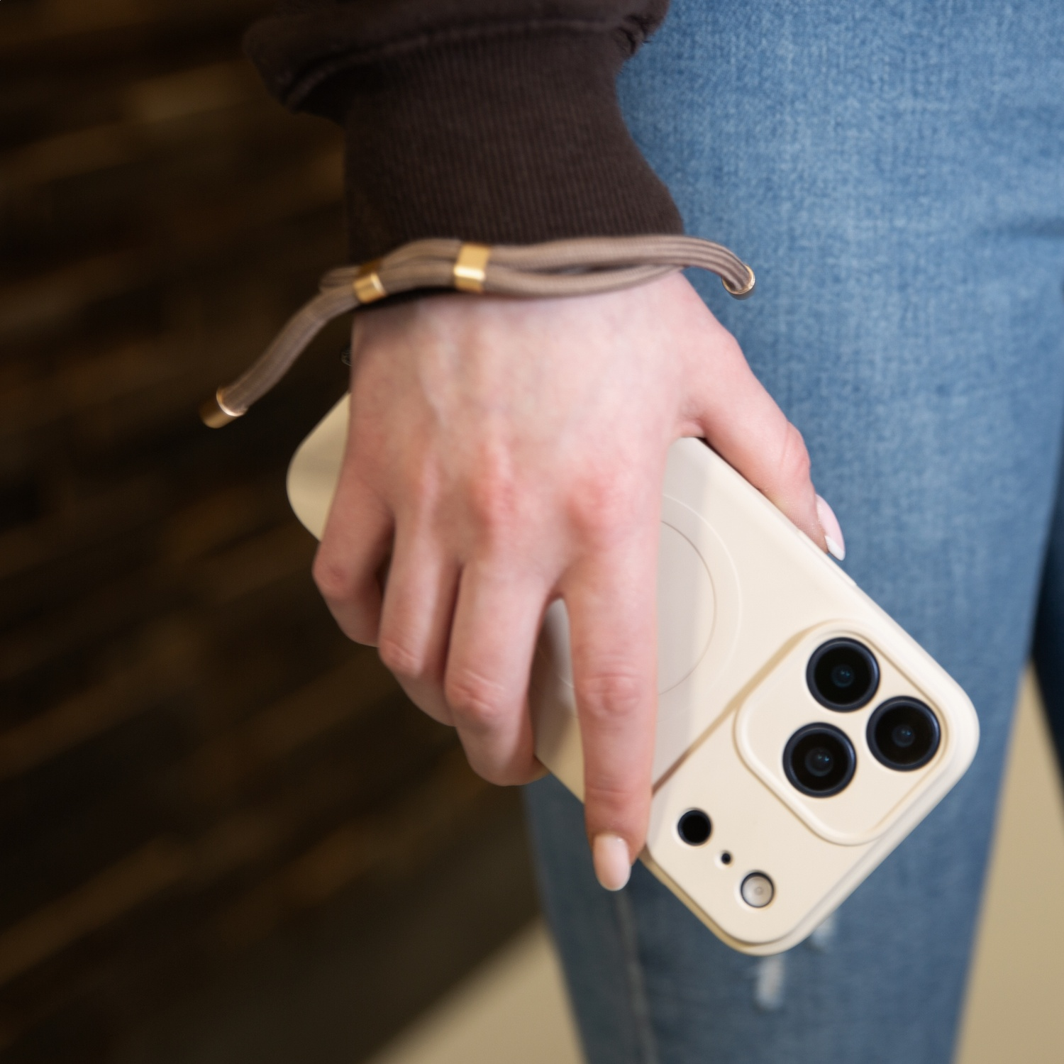 A person in jeans and a brown hoodie holds a beige phone case with three cameras.