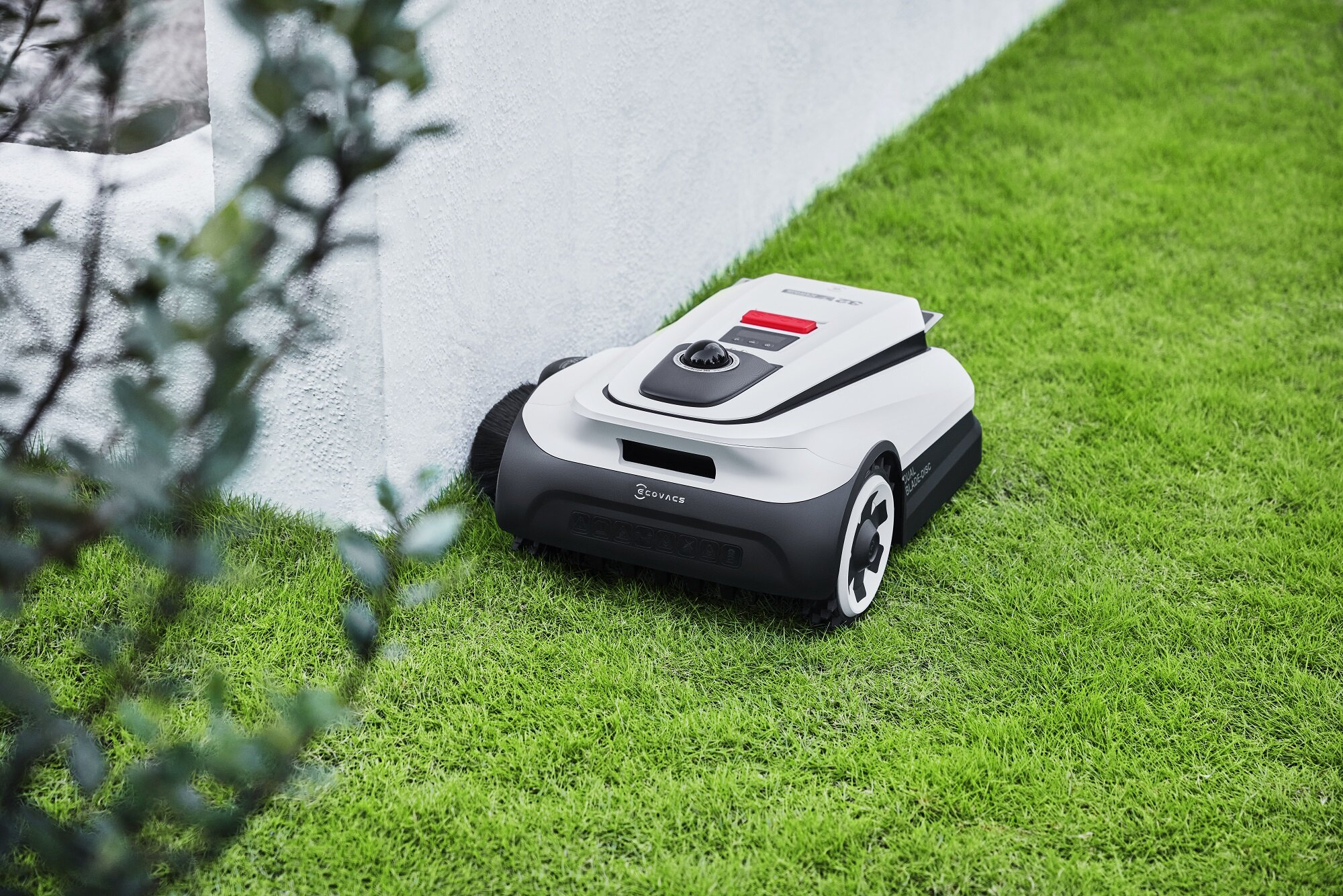 Biały robot koszący z czarną podstawą kosi zielony trawnik obok białej ściany.