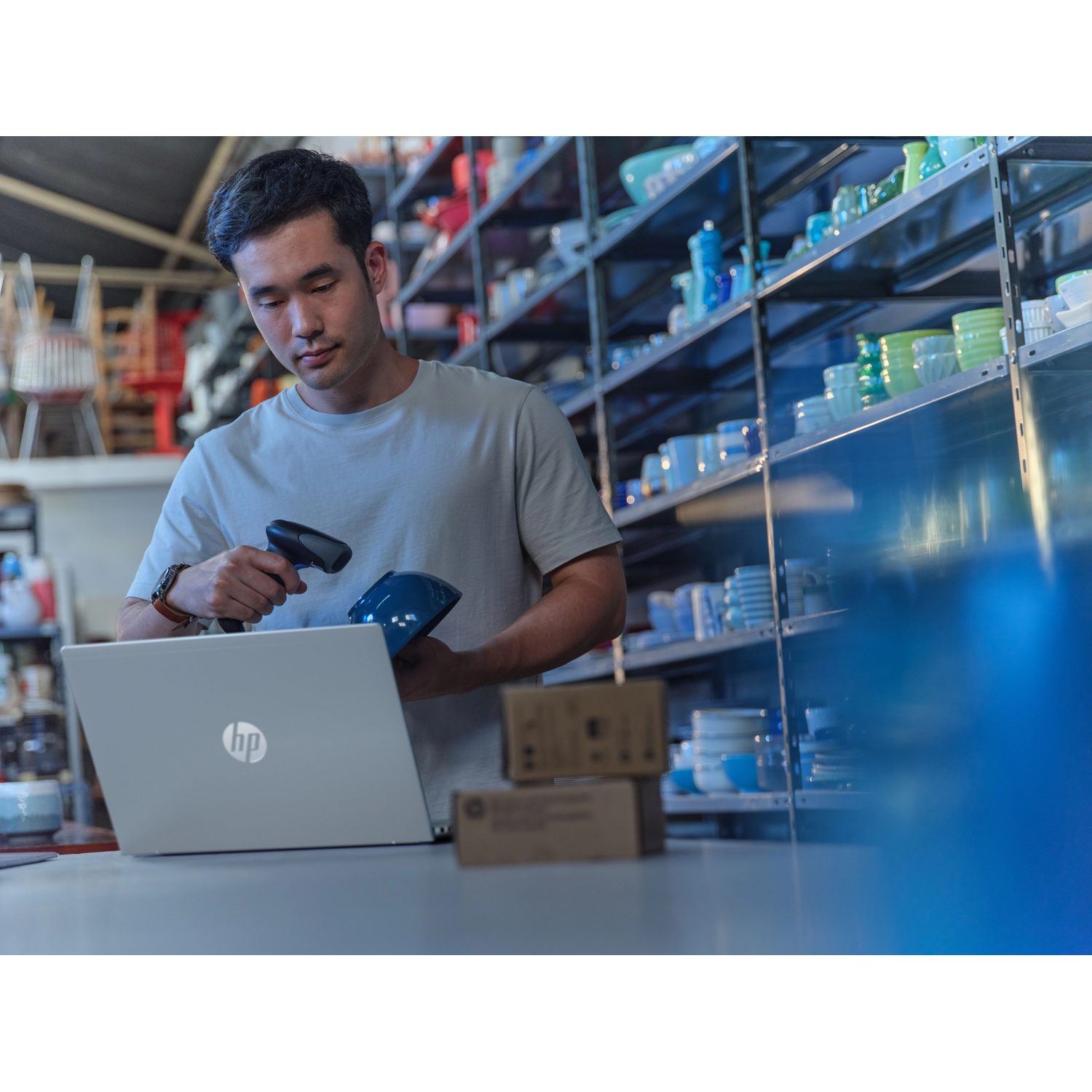 Een man scant een blauw object voor een laptop in een magazijn met schappen vol servies.