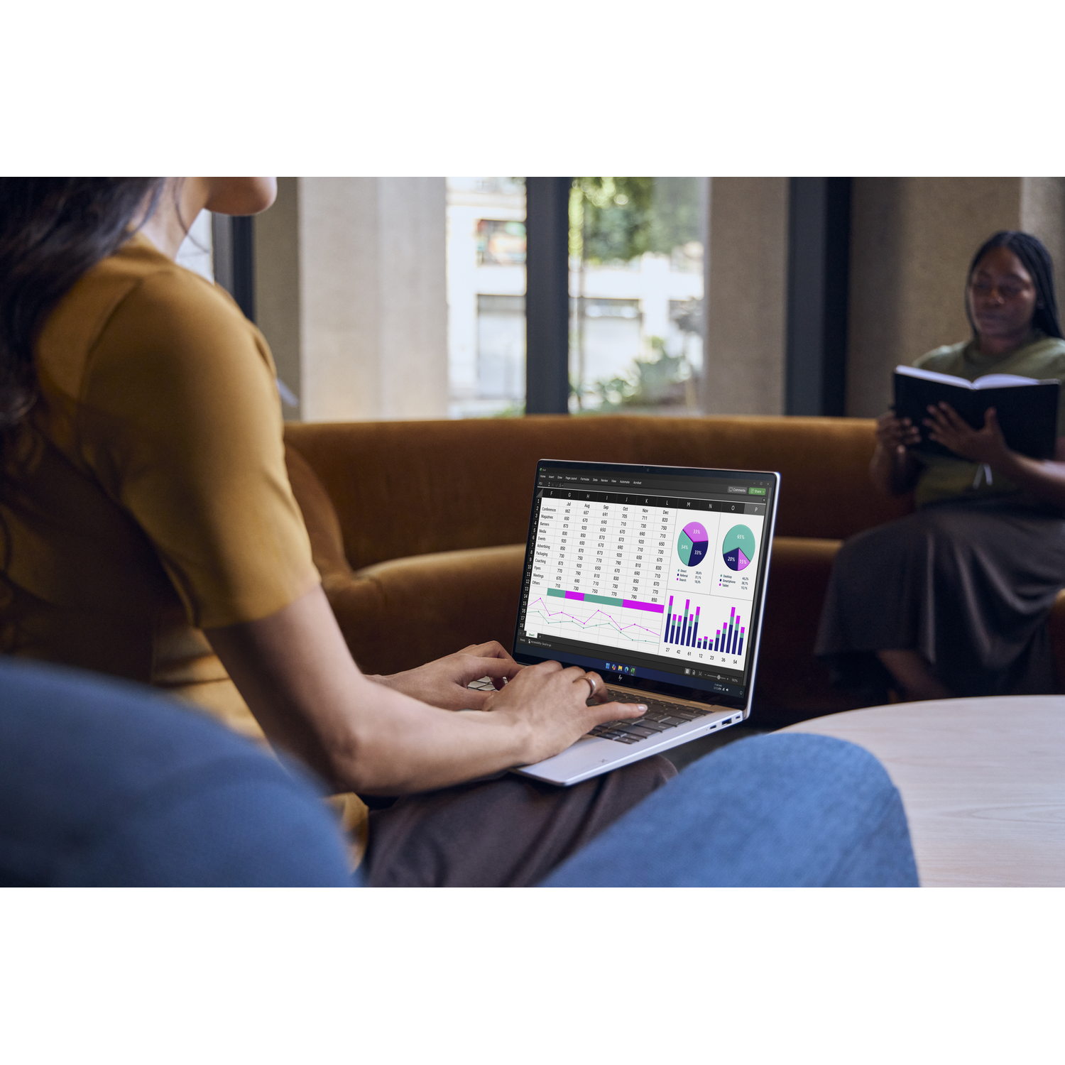 Een vrouw typt op een laptop met financiële grafieken. Een andere vrouw leest op de achtergrond.