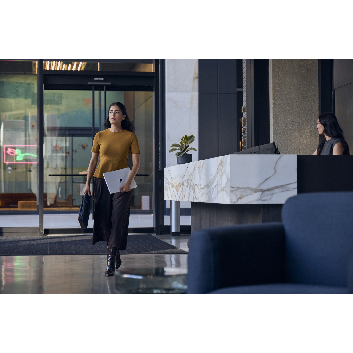 Een vrouw met laptop en tas loopt door een lobby. Een receptioniste is op de achtergrond.
