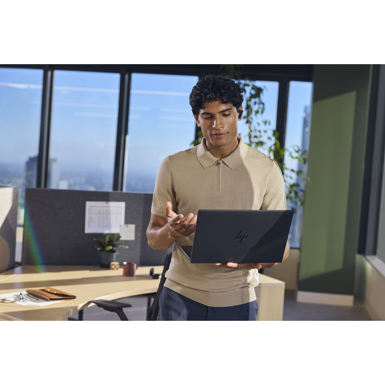 Een man staat in een kantoor en houdt een laptop vast. Hij draagt een beige poloshirt.