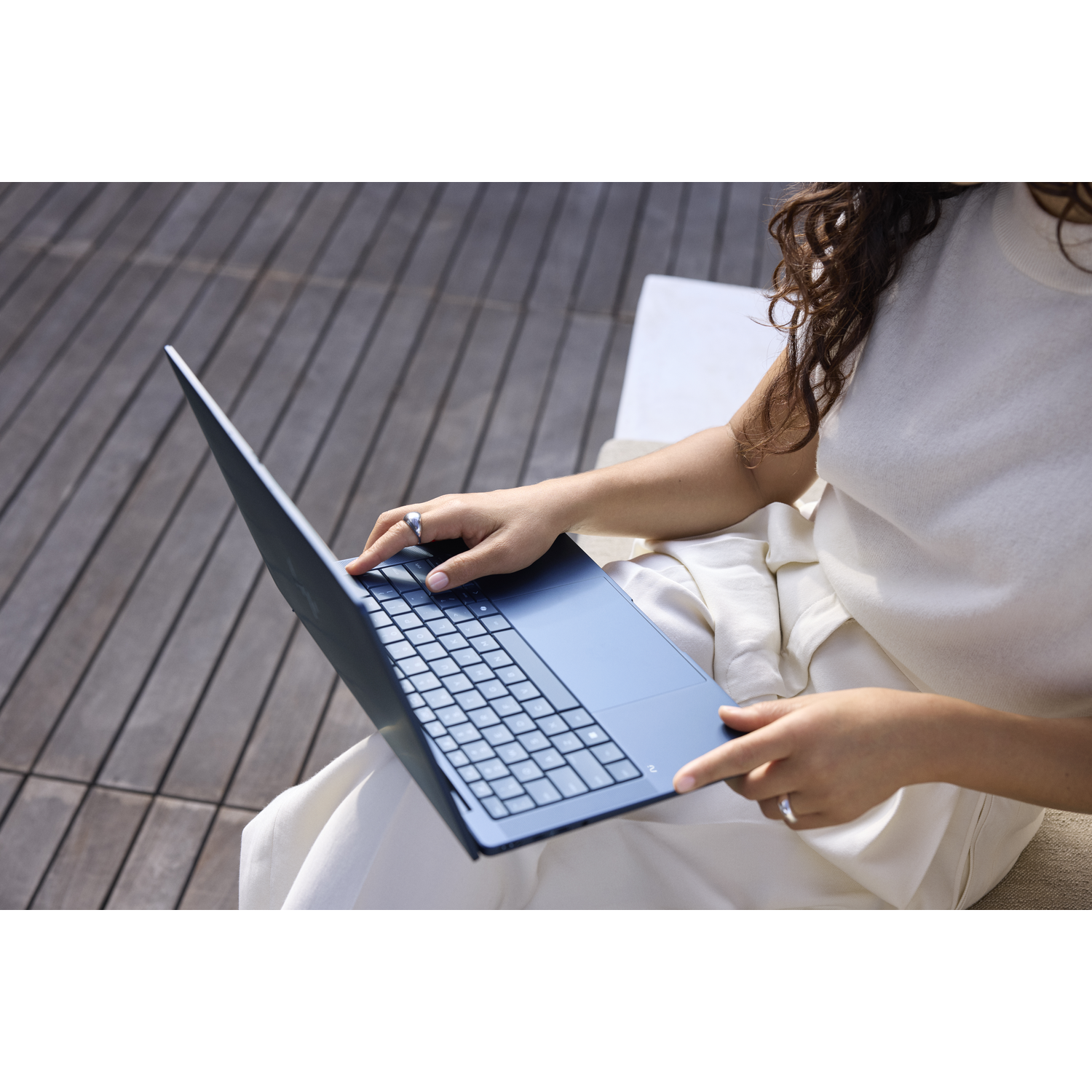 Een vrouw met donker krullend haar gebruikt een blauwe laptop op een houten dek.