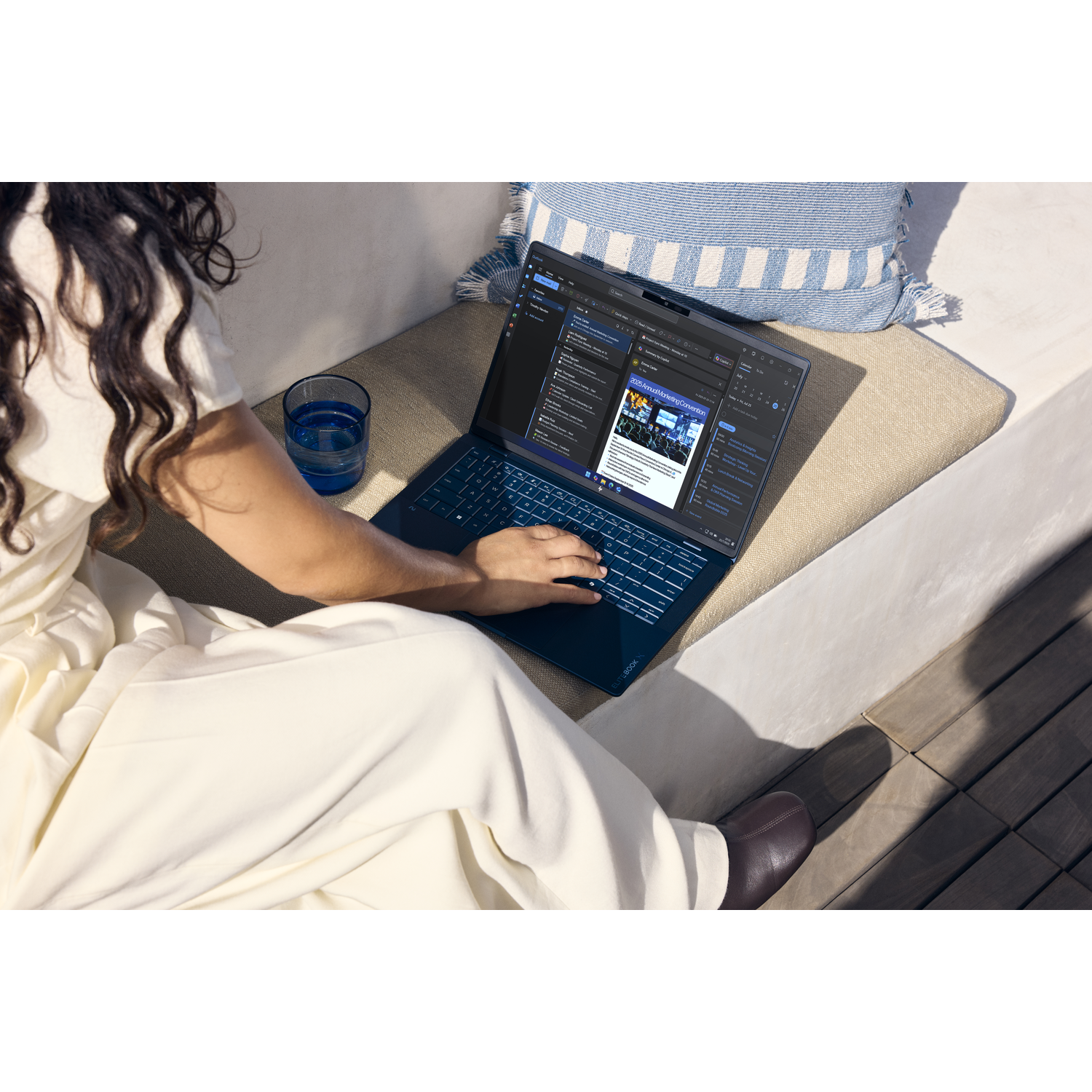 Een persoon typt op een donkere laptop op een bankje. Een glas water staat ernaast.