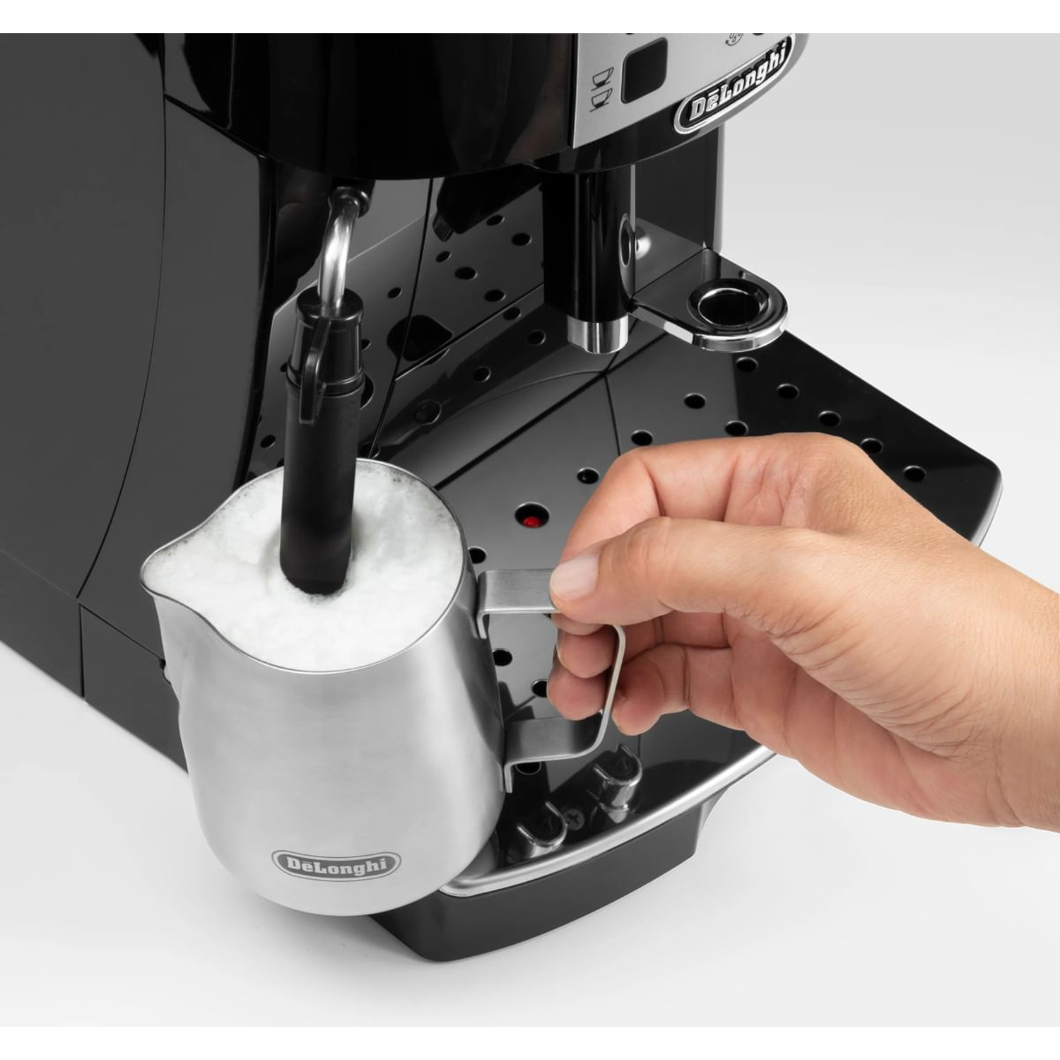 Een hand houdt een melkopschuimkan met schuim vast naast een koffiezetapparaat.