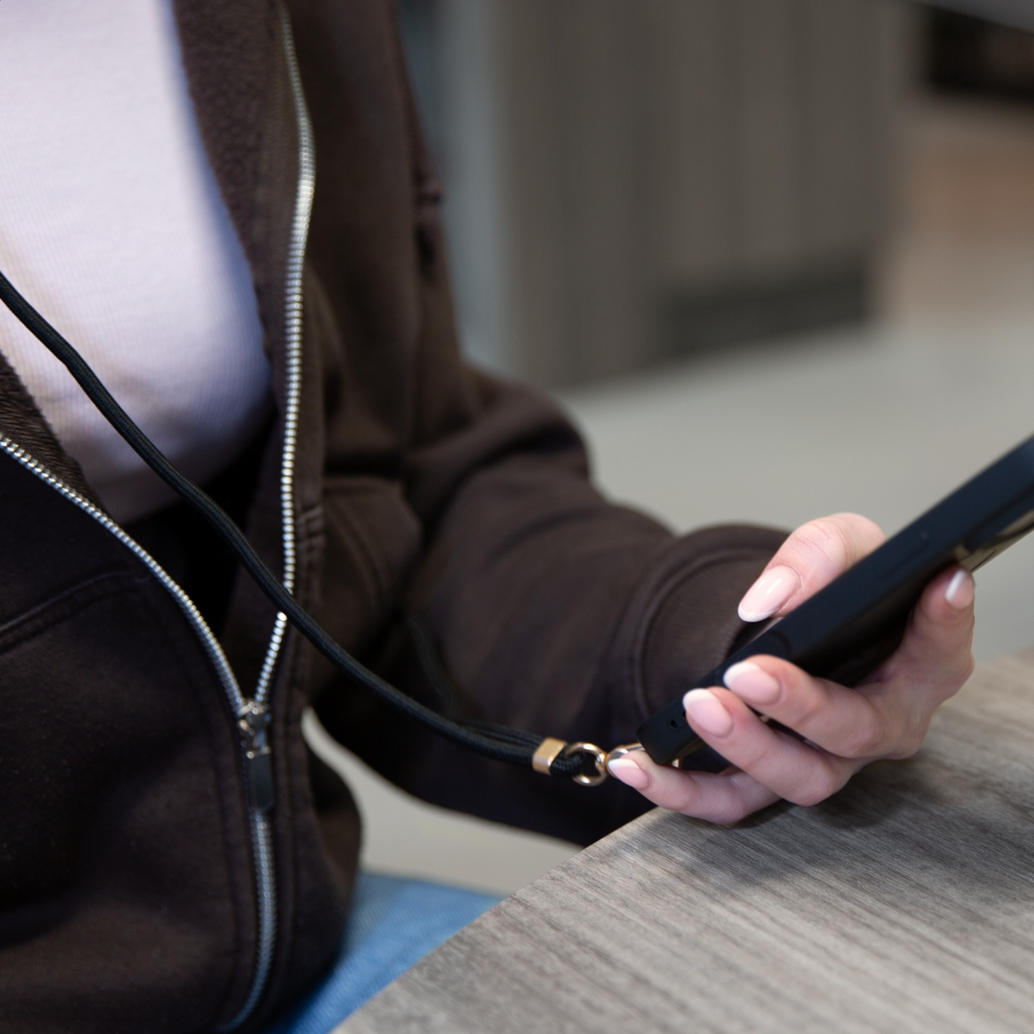 A woman holds a smartphone with a lanyard, seated at a wooden table.