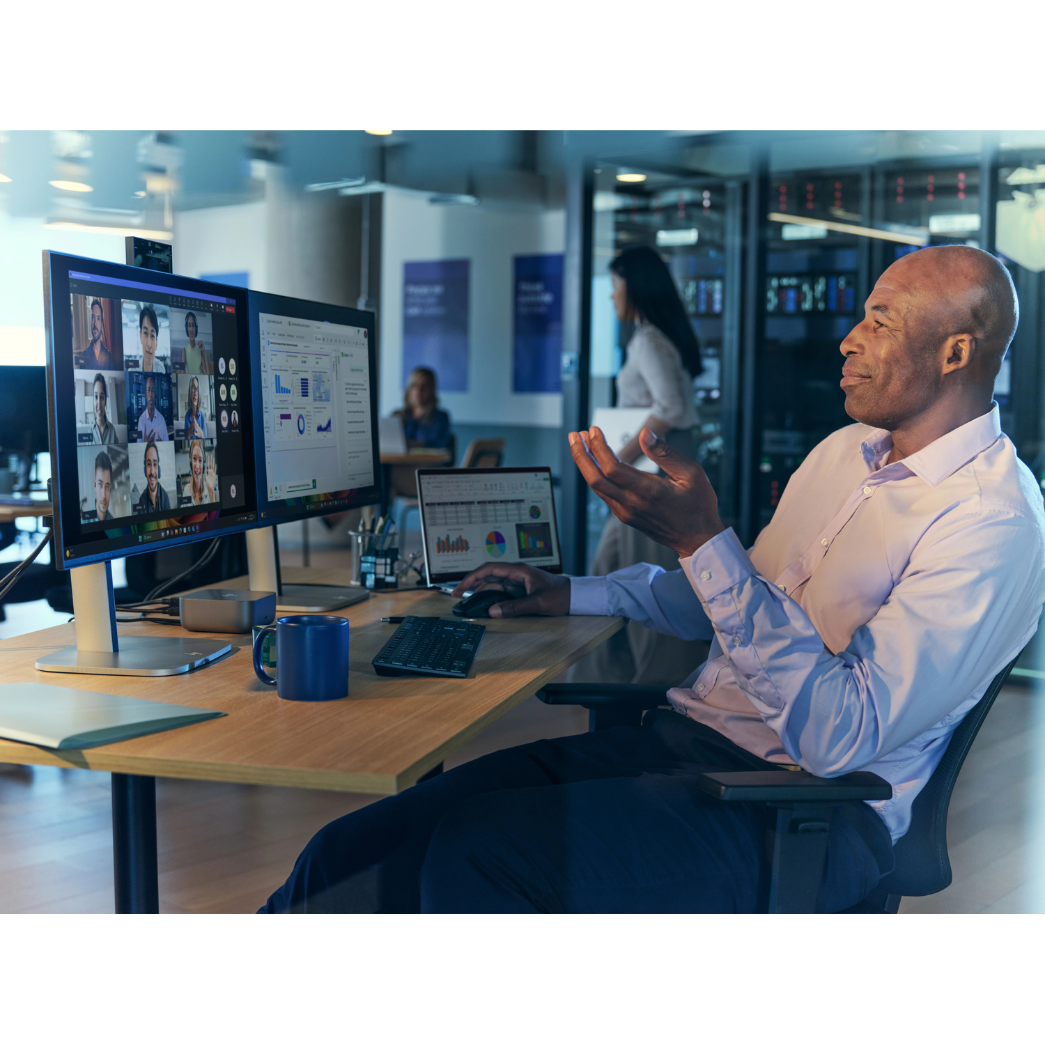 Man in overhemd in videogesprek op twee monitoren, serverruimtes op de achtergrond.