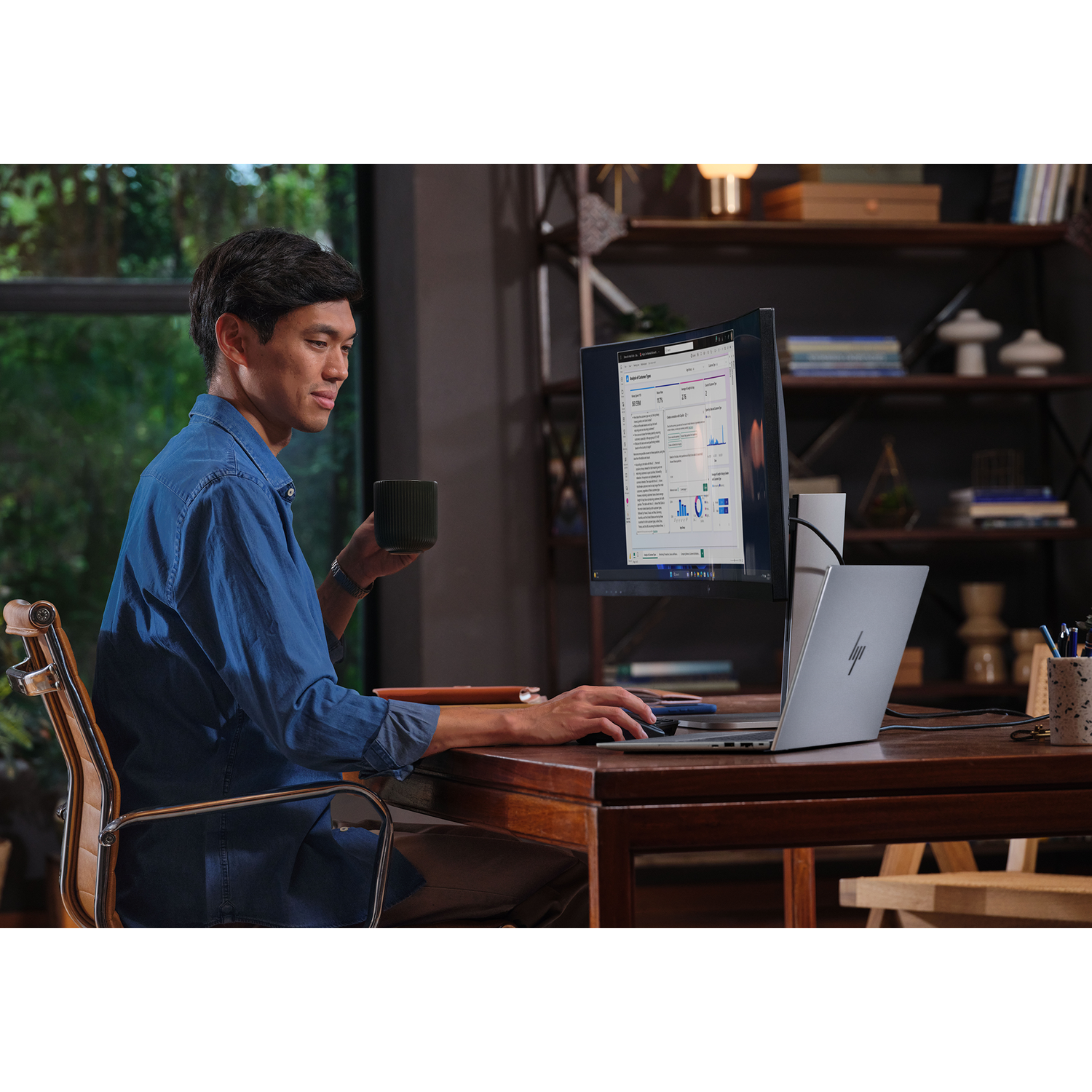 Man in blauw shirt aan bureau met laptop en monitor, houdt mok vast.