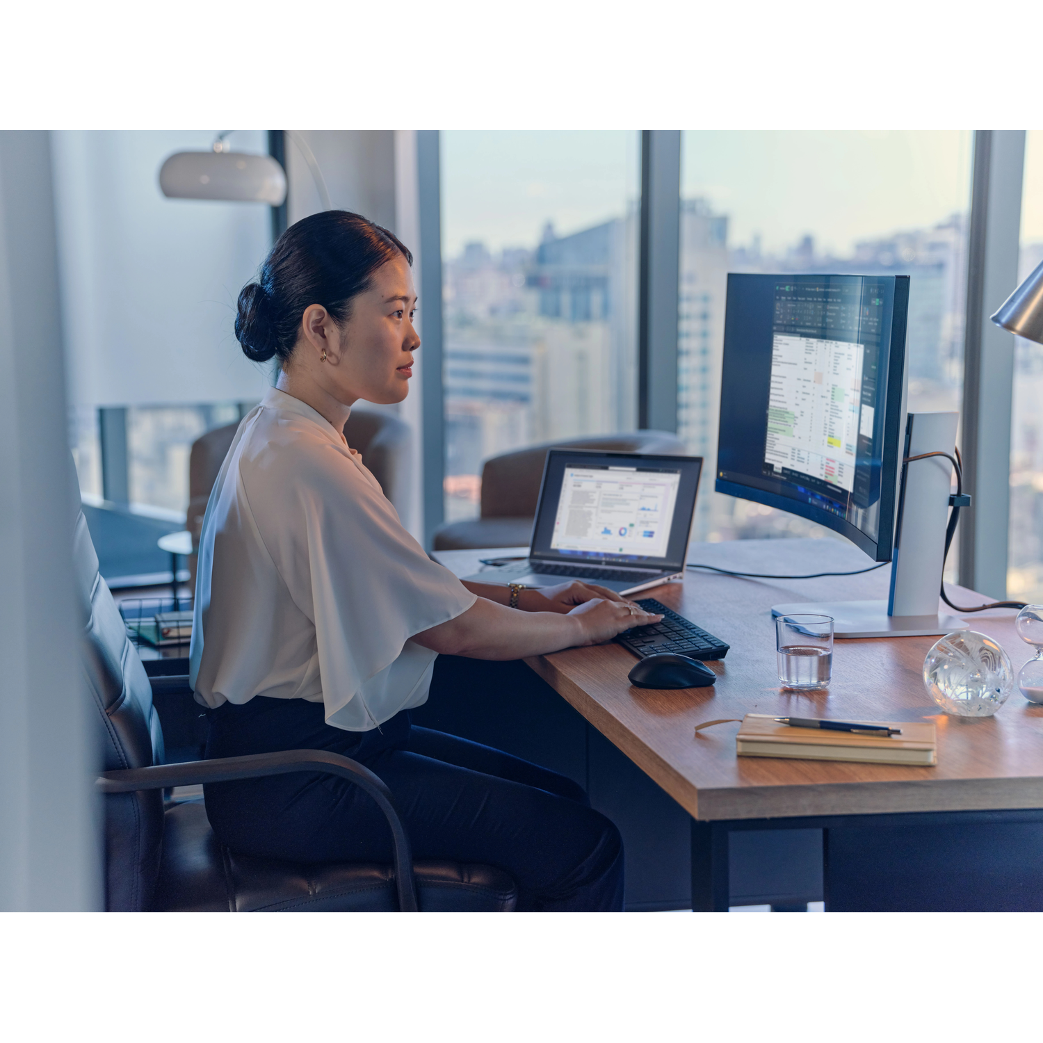 Vrouw in wit shirt werkt op computer aan bureau met uitzicht op de stad.
