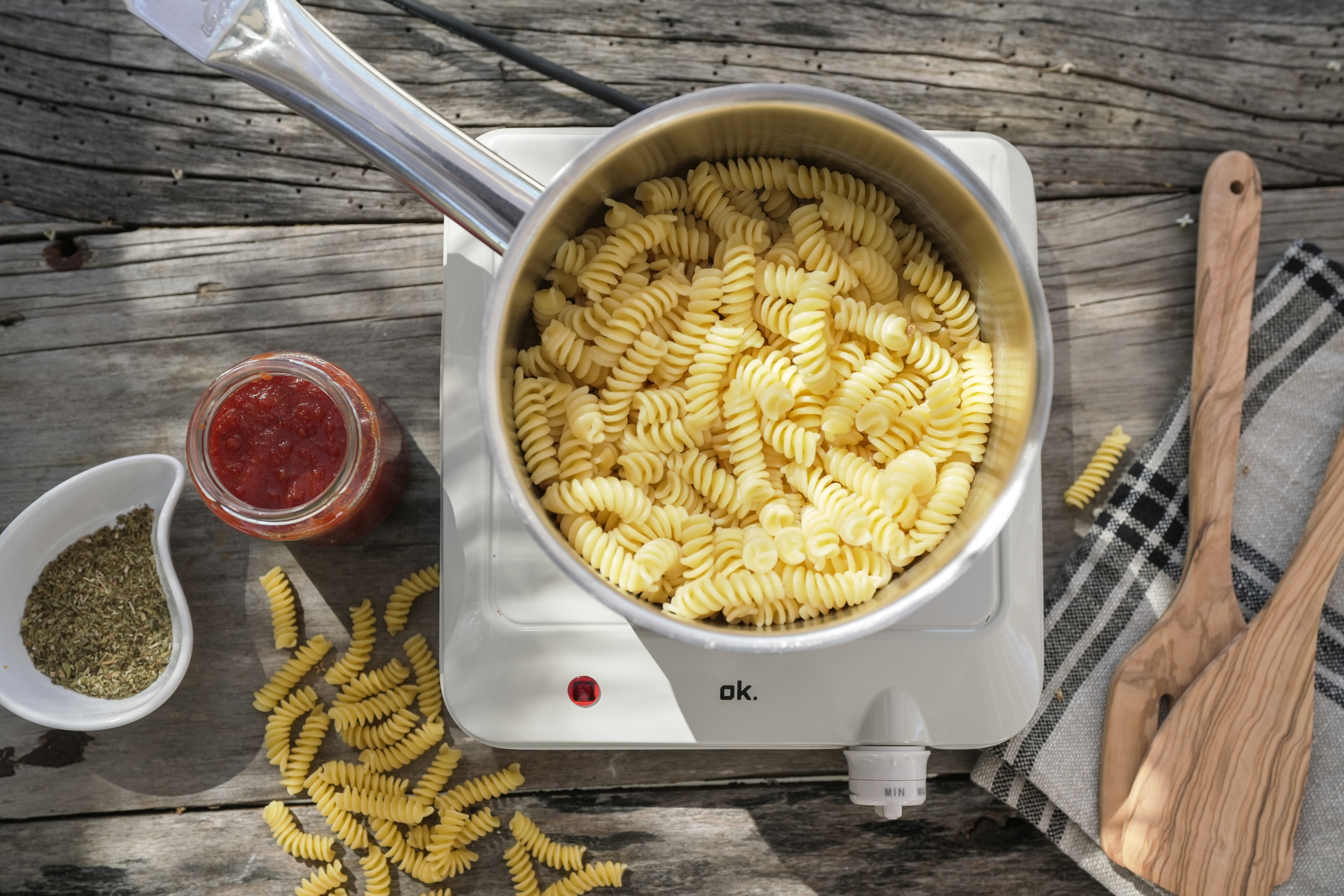 Une casserole de pâtes fusilli sur une plaque chauffante, à côté d'un bocal de sauce tomate et d'herbes.