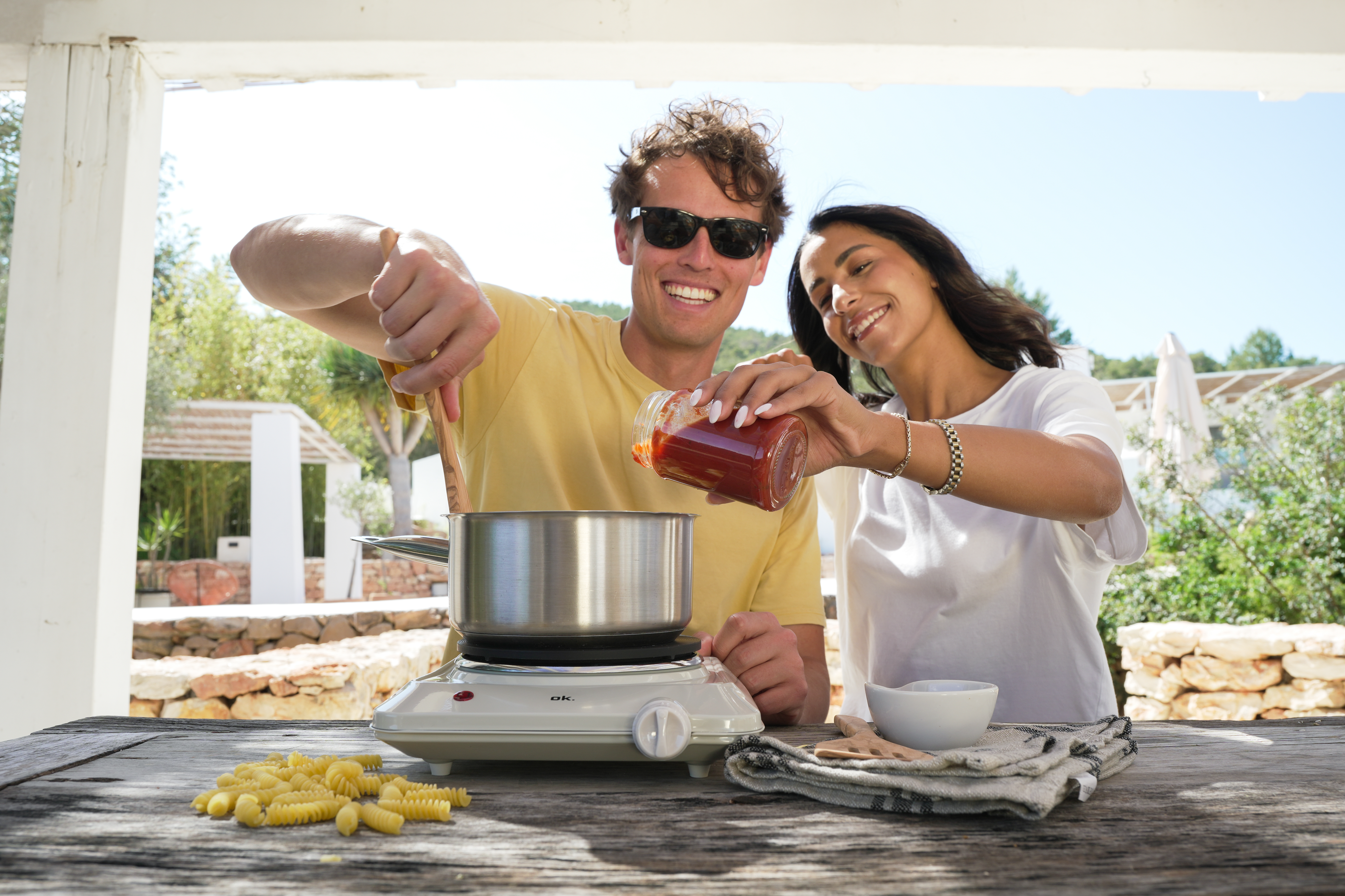 Homme verse sauce tomate dans casserole, femme sourit. Pâtes sur table.