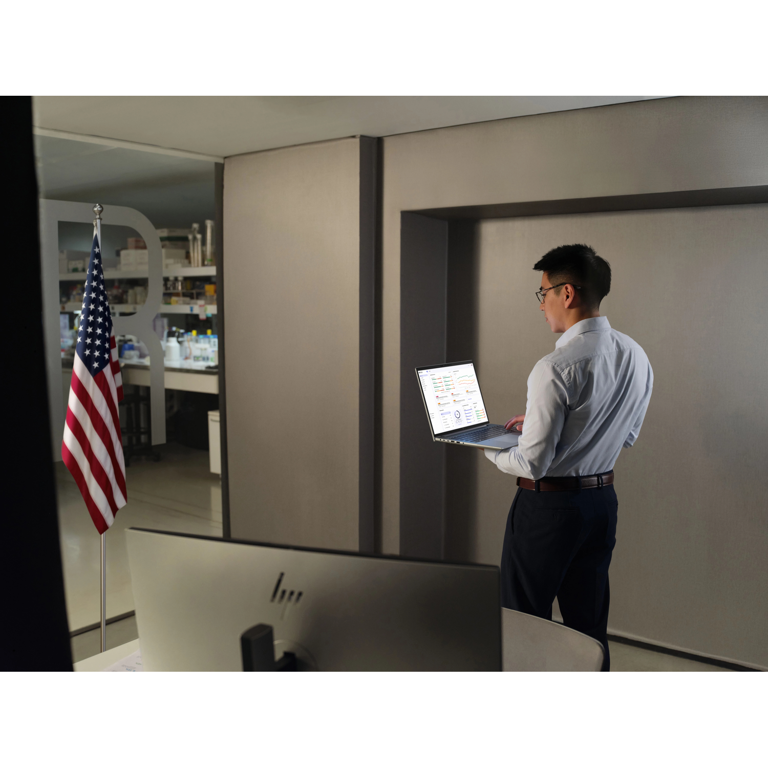 Man met laptop in kantoor met Amerikaanse vlag en monitor. Grafieken zichtbaar.