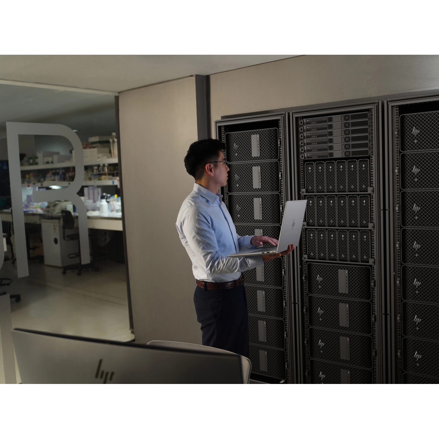 Man in lichtblauw shirt werkt op laptop voor serverracks. Laboratorium zichtbaar op achtergrond.