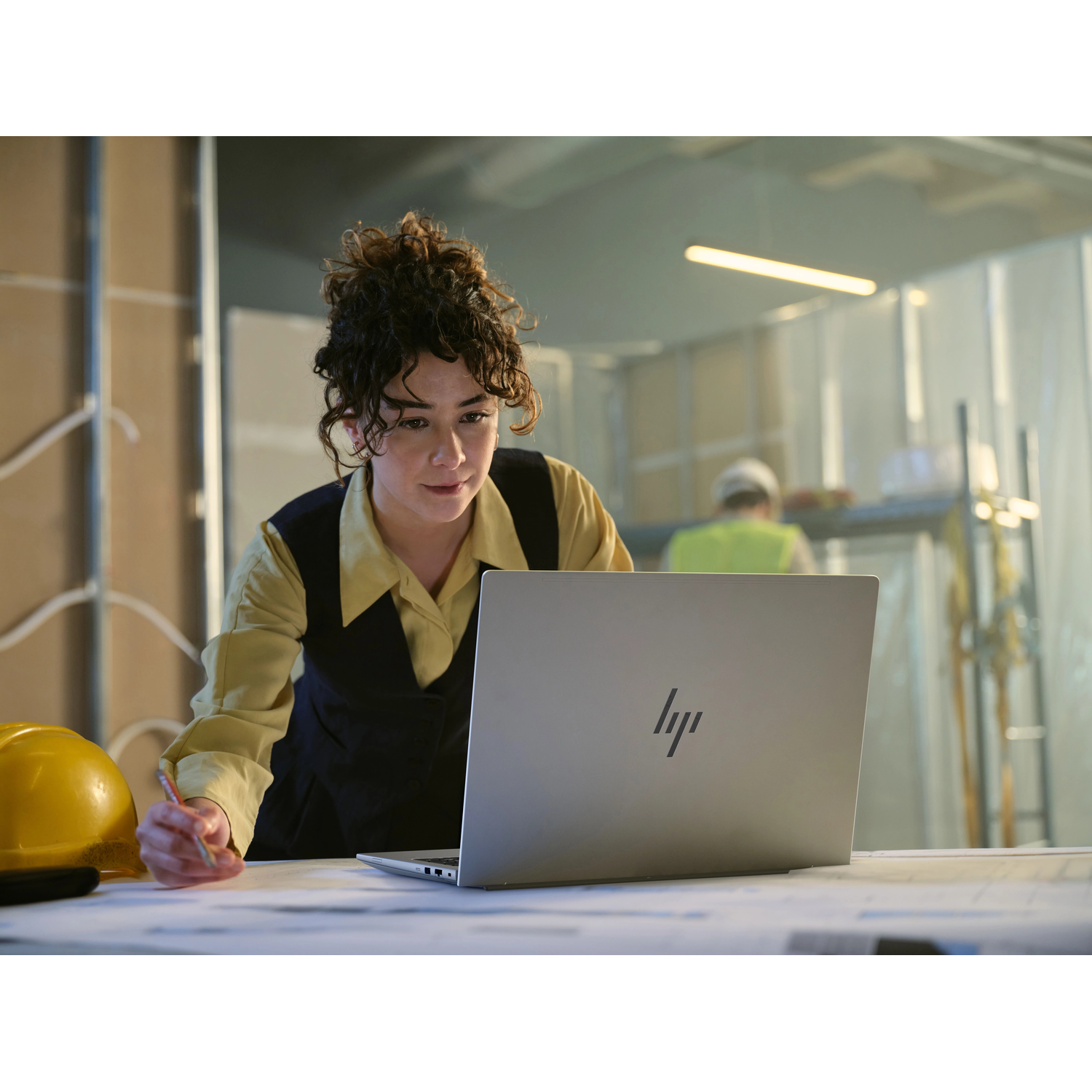 Een vrouw met laptop en gele helm op bouwplaats.