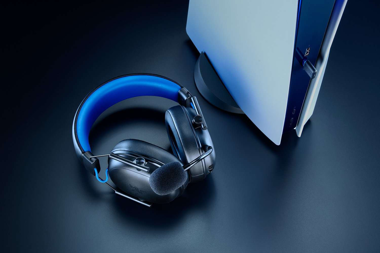 Auriculares negros con diadema azul junto a una consola de juegos blanca sobre fondo oscuro.