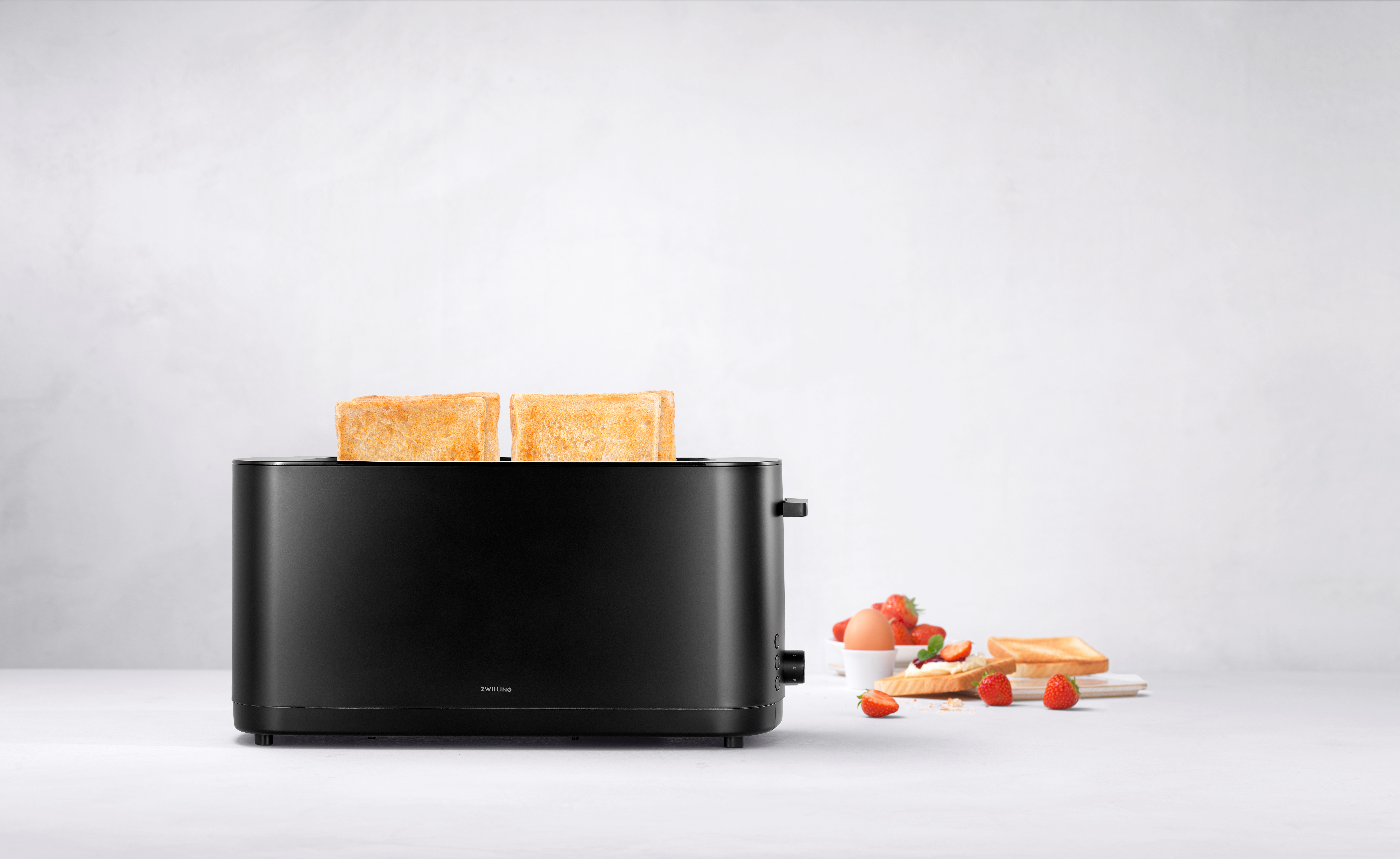 Ein schwarzer Toaster mit zwei Scheiben Toast, daneben Toast mit Erdbeeren und Ei.