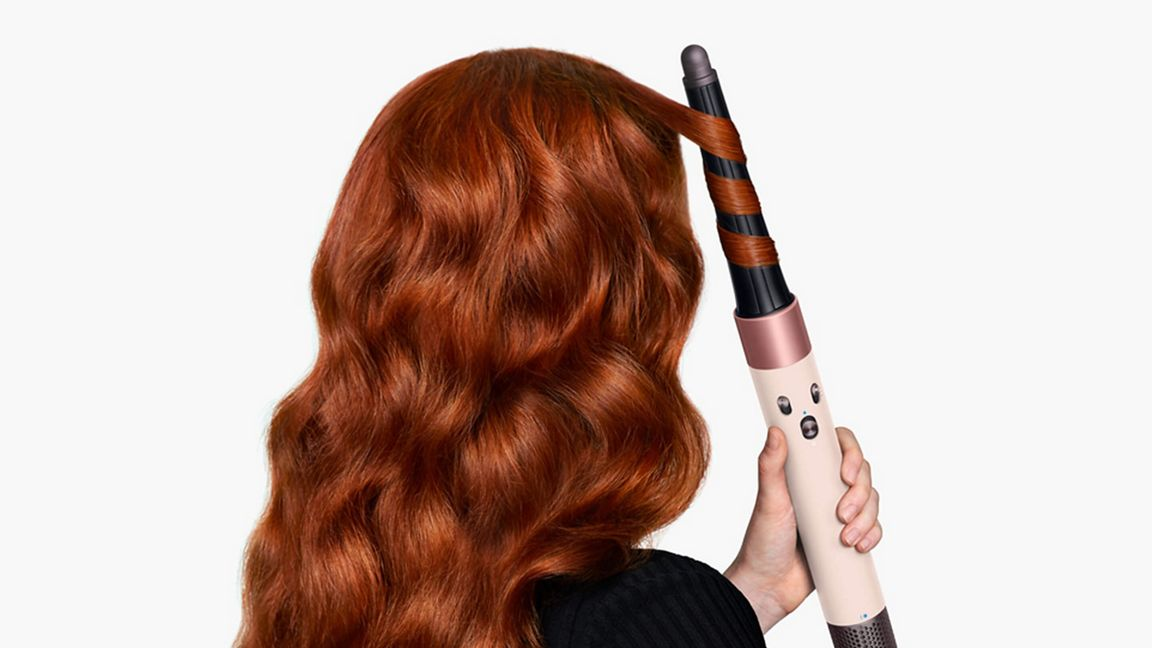 Una persona con lunghi capelli ondulati color rame usa un arricciacapelli. L'arricciacapelli è beige con accenti oro rosa.
