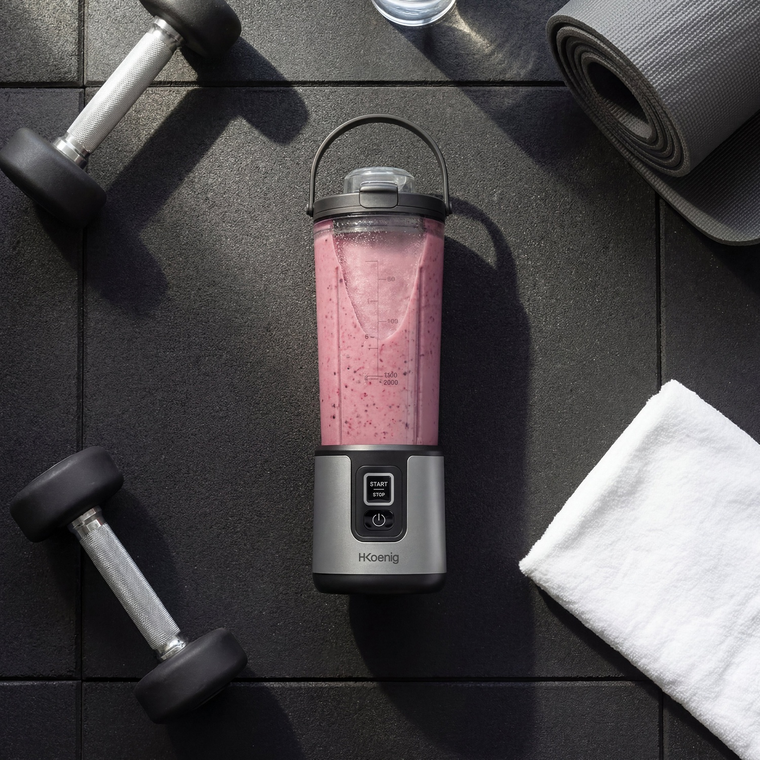 Un frullatore portatile con frullato di bacche, manubri, tappetino yoga e asciugamano su pavimento scuro.