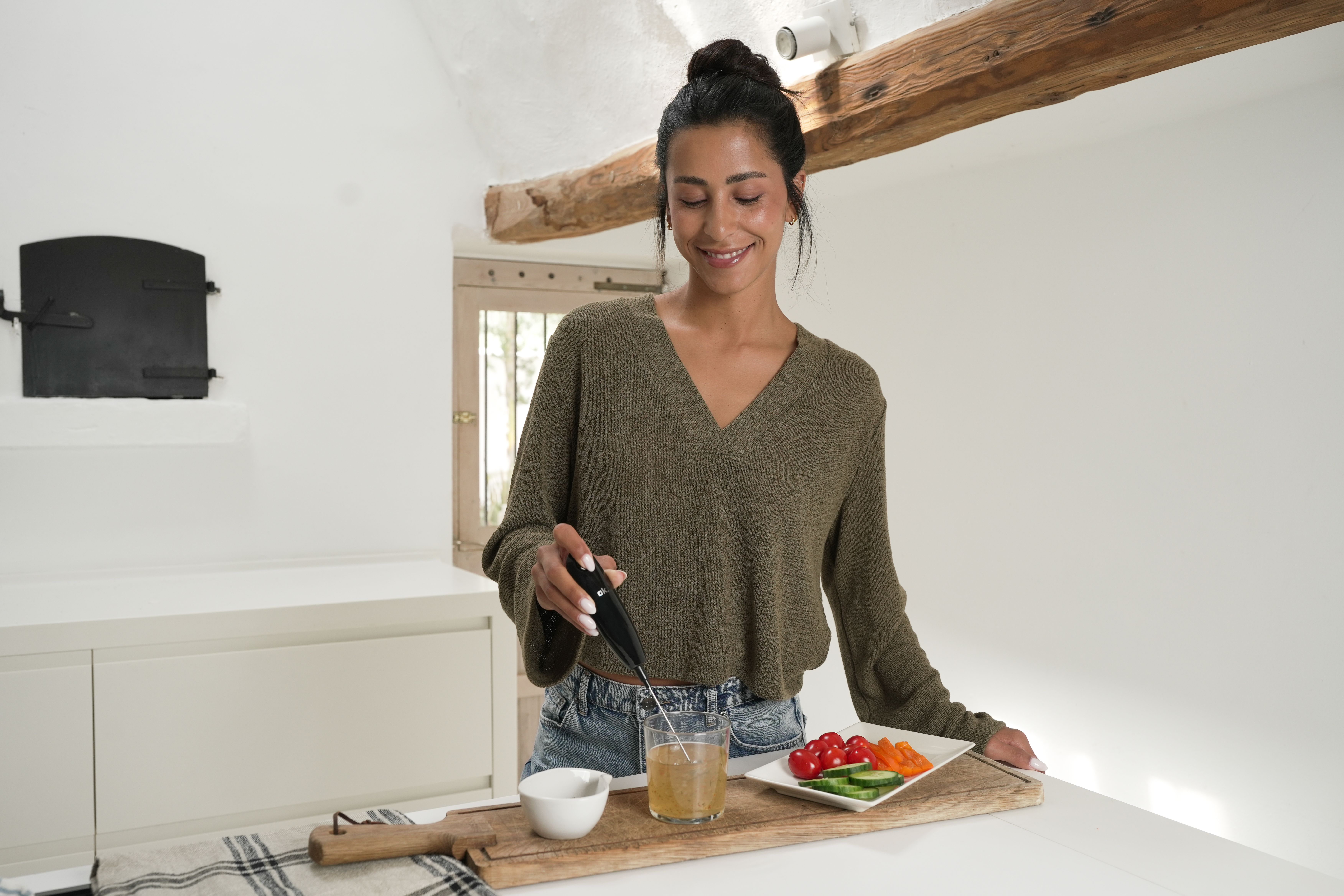Une femme utilise un mixeur plongeant dans un verre, avec des légumes dans une assiette à proximité.
