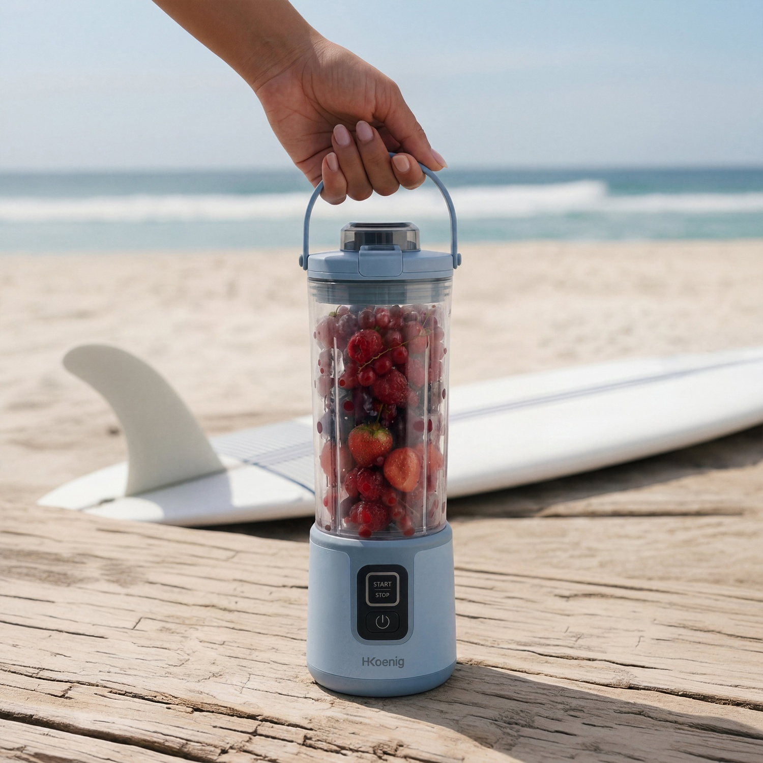 Una mano tiene un frullatore con bacche su una spiaggia, accanto a una tavola da surf.
