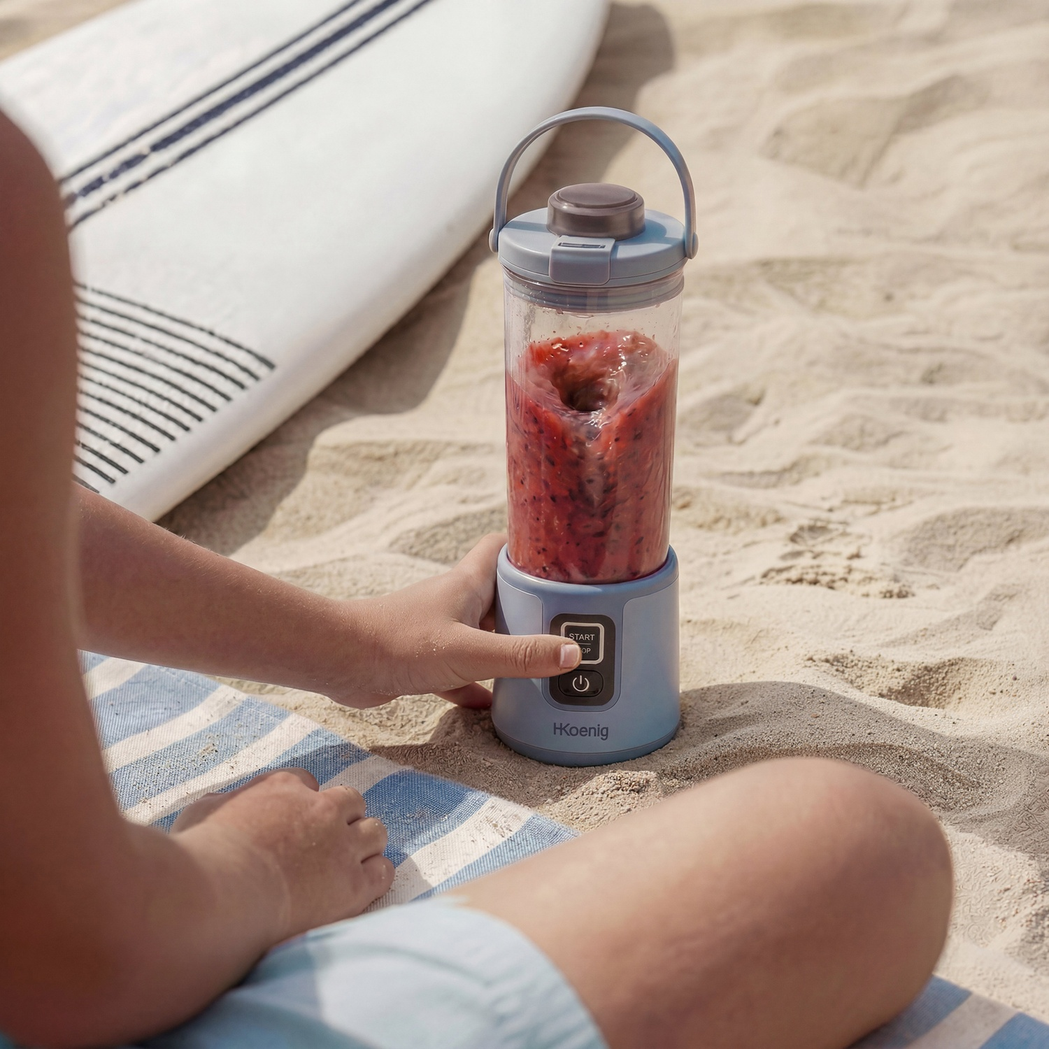 Un frullatore portatile sulla sabbia, accanto a una tavola da surf, con una mano che lo aziona.