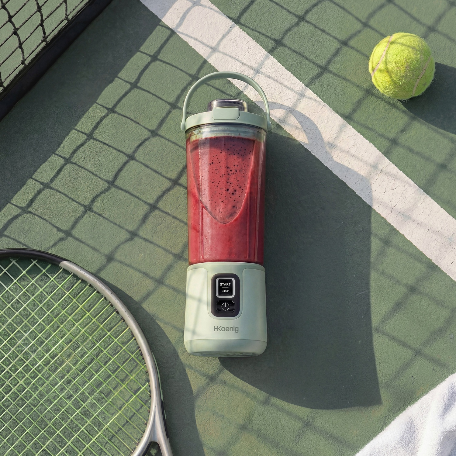 Un frullatore verde con frullato rosso è su un campo da tennis accanto a una racchetta e una palla.