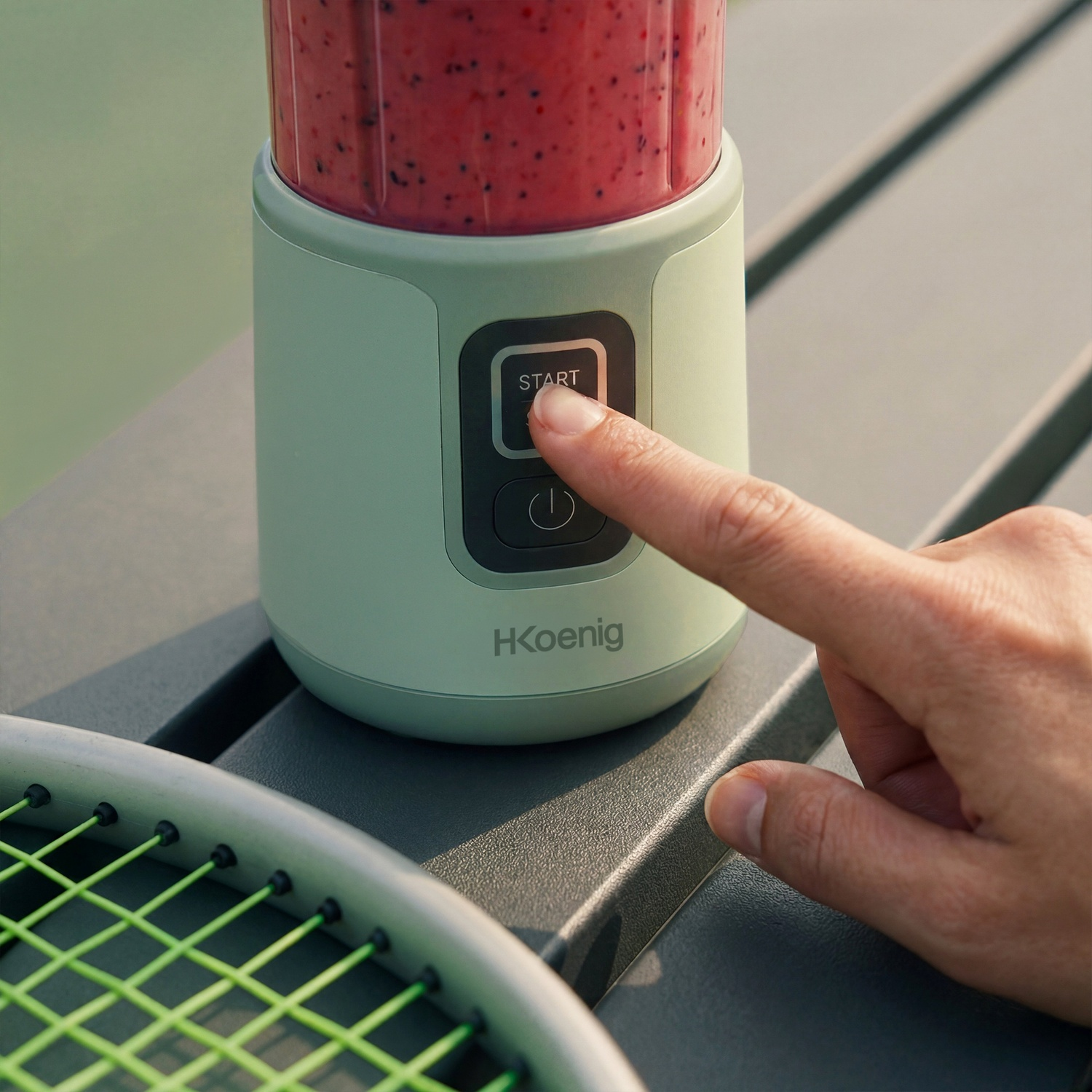 Un dito preme il pulsante di avvio di un frullatore verde con frullato rosso.