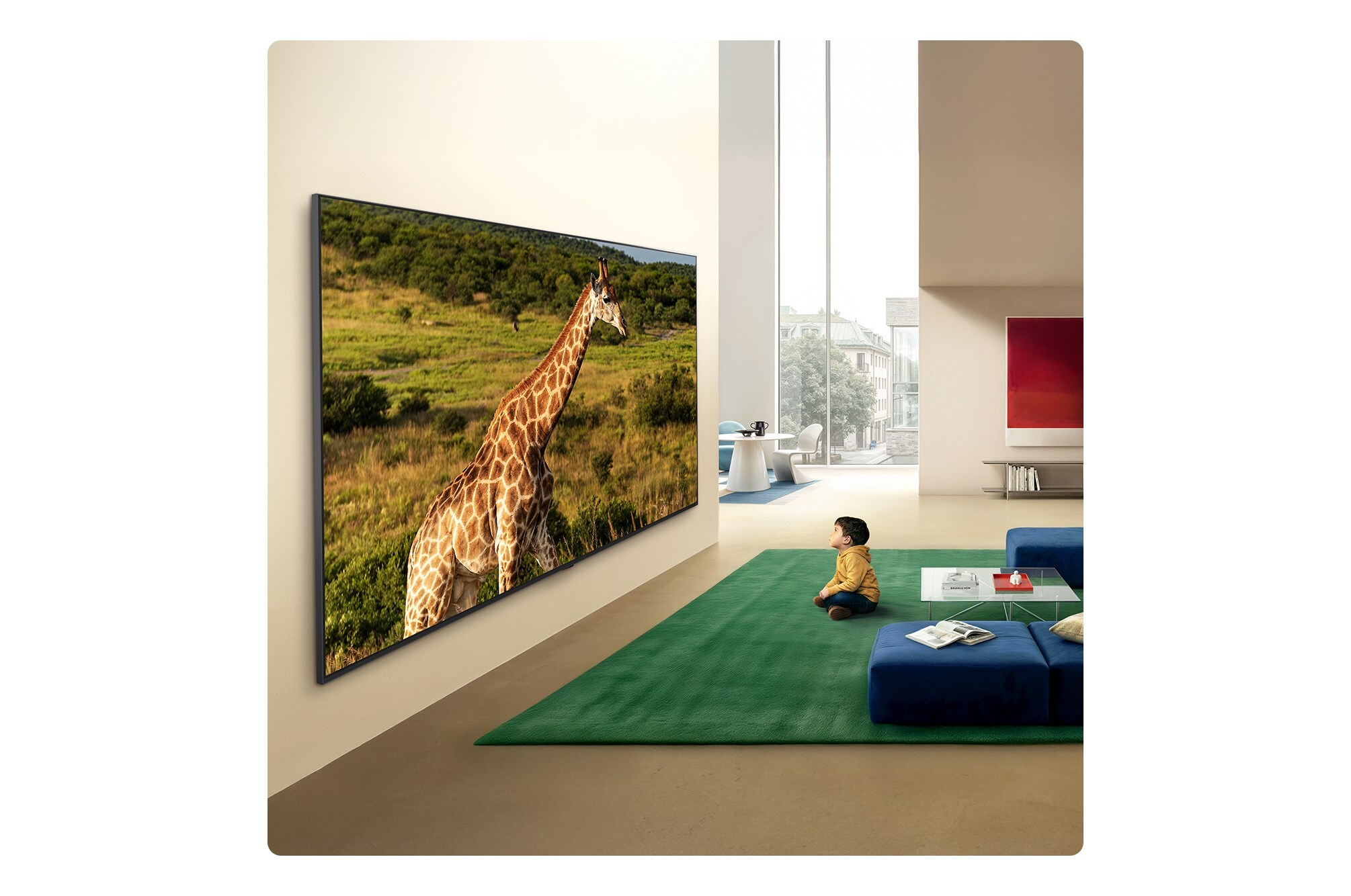 Un garçon assis sur un tapis vert regarde un grand écran de télévision affichant une girafe.