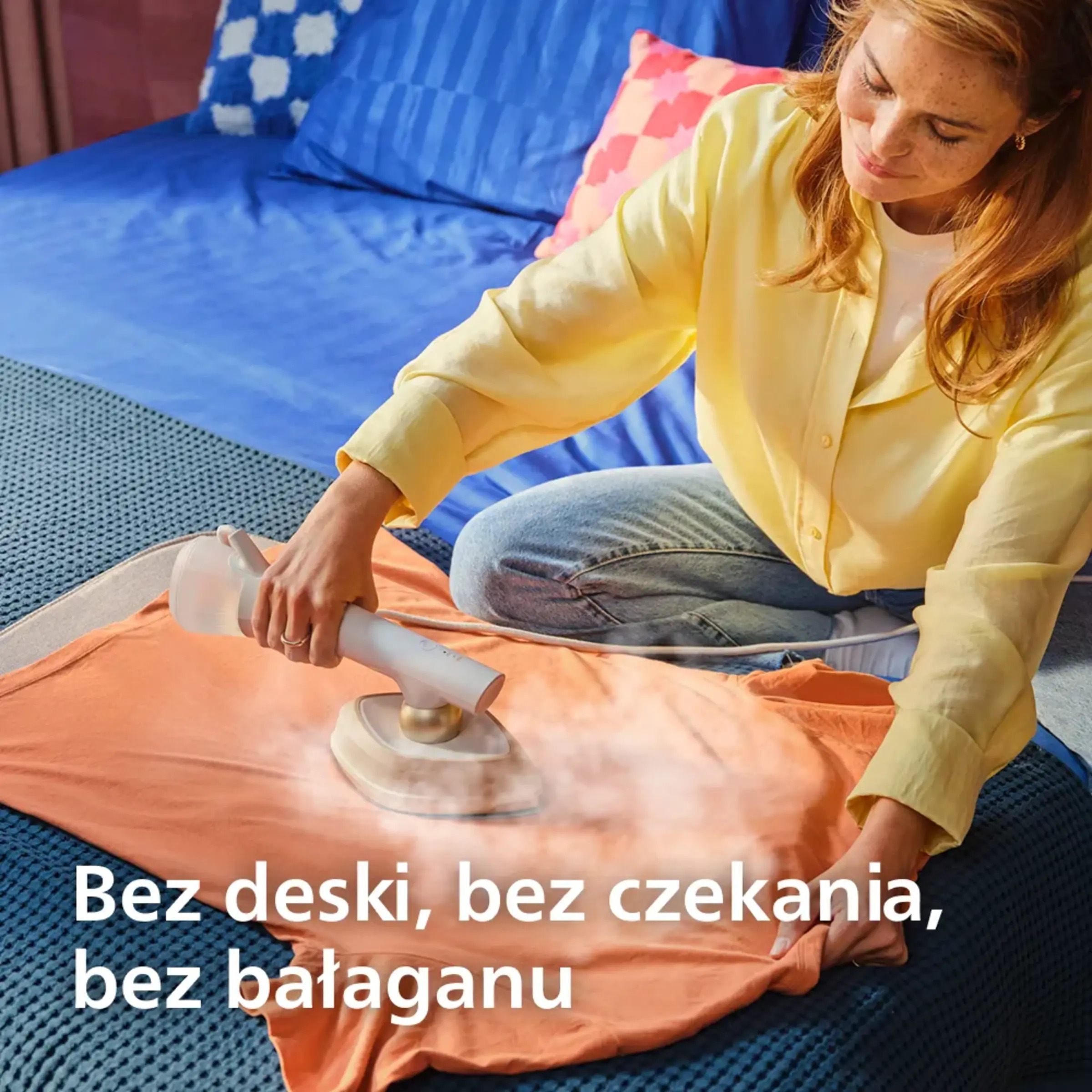 Kobieta prasuje pomarańczową koszulkę ręcznym parownikiem na łóżku.