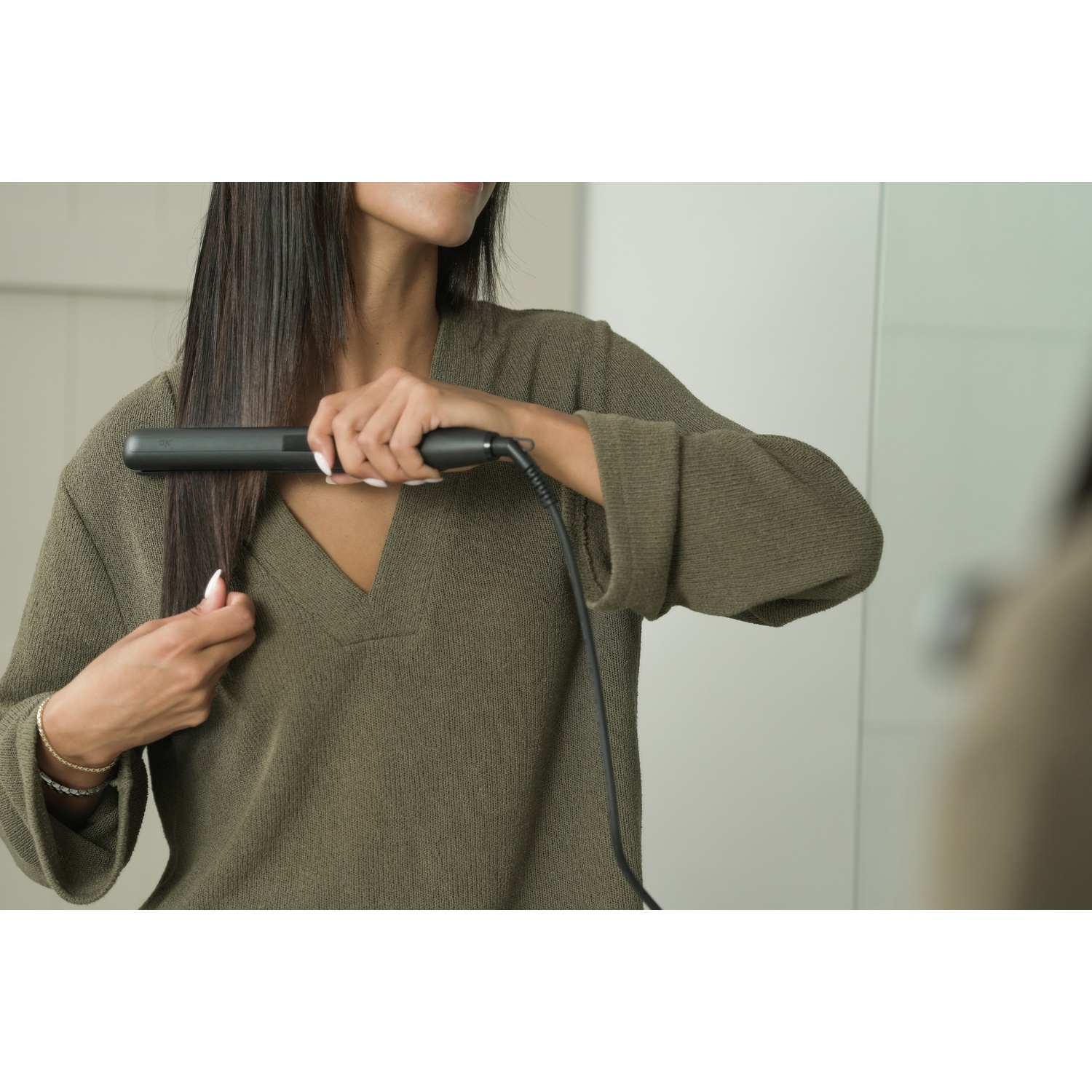 Una donna si liscia i lunghi capelli scuri con una piastra nera. Indossa un maglione verde oliva.