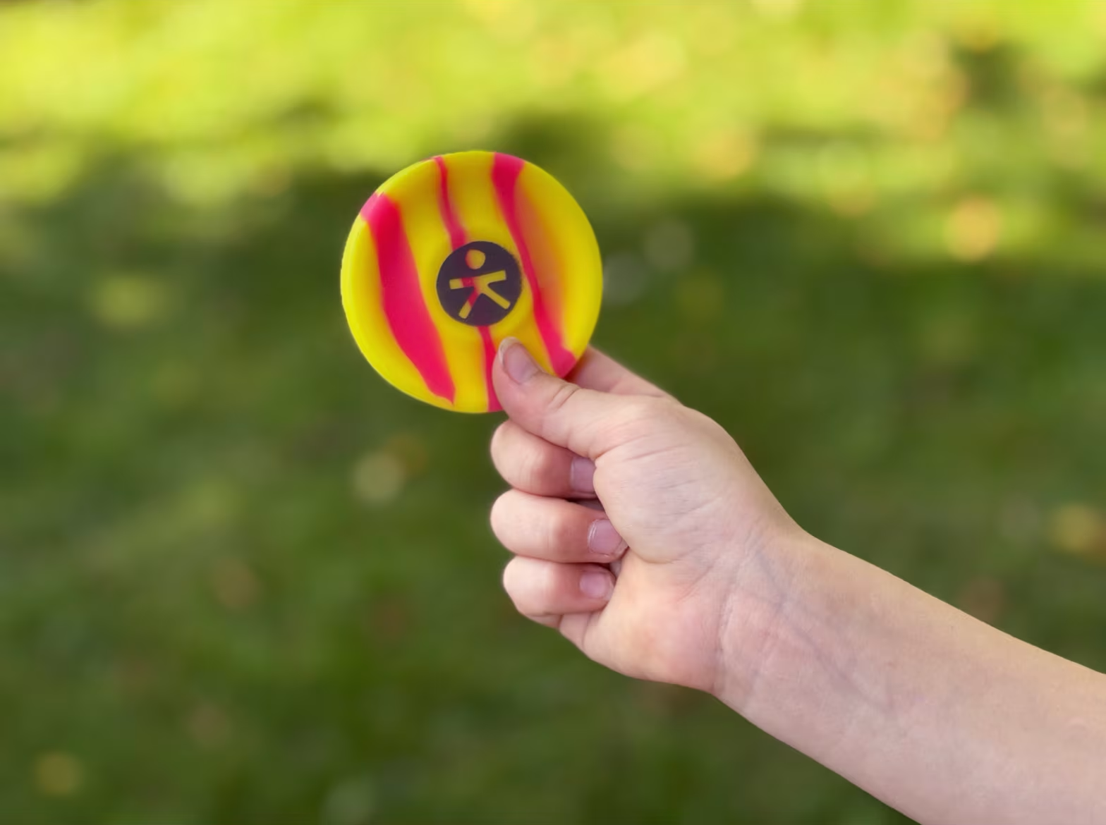 Egy kéz sárga és rózsaszín csíkos korongot tart, fekete szimbólummal.