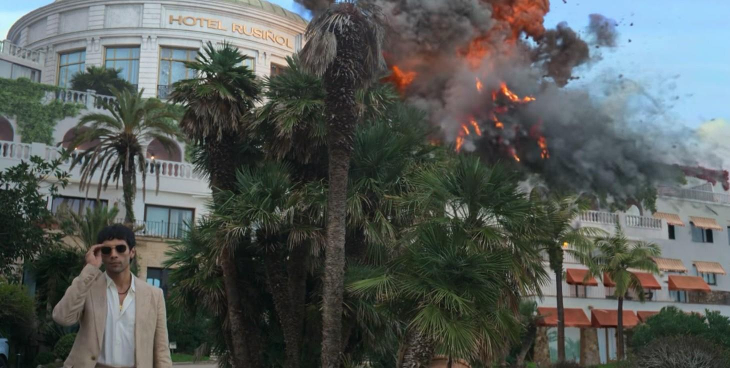 Ein Mann in einem beigen Anzug vor einem Hotel, während eine Explosion Feuer und Rauch verursacht.