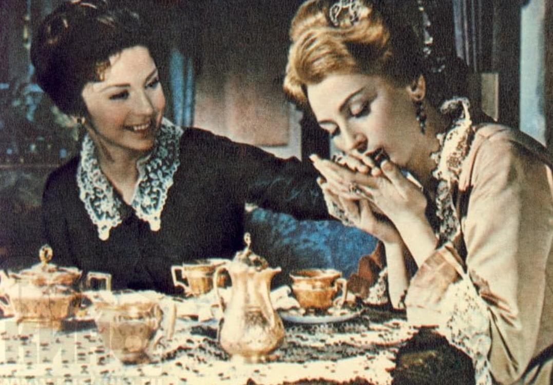 Zwei Frauen an einem Tisch mit goldenem Geschirr, eine küsst die Hand der anderen.