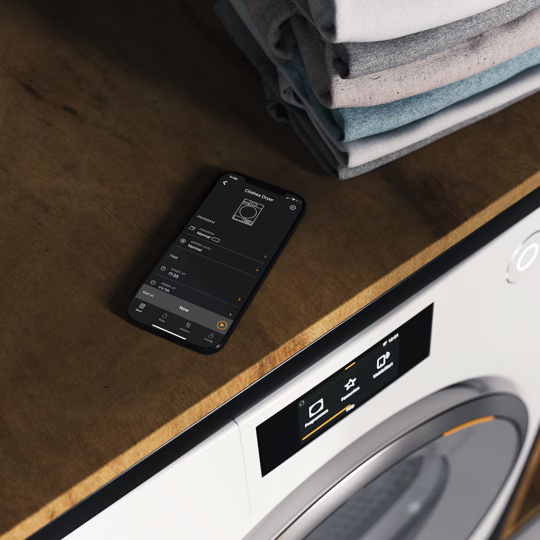 Smartfon z aplikacją suszarki leży na drewnianej powierzchni obok pralki.