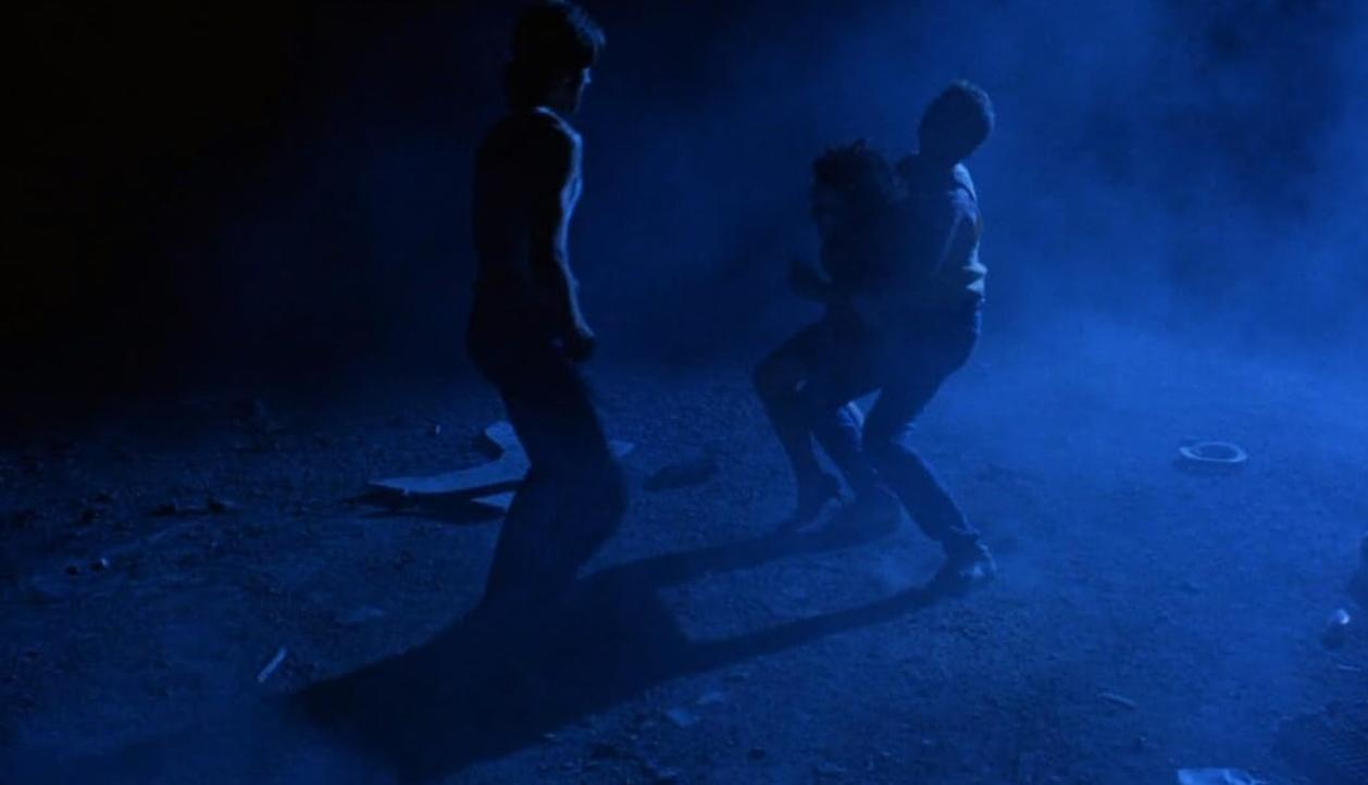 Zwei Männer kämpfen in blauer Nacht. Einer steht, der andere ringt.