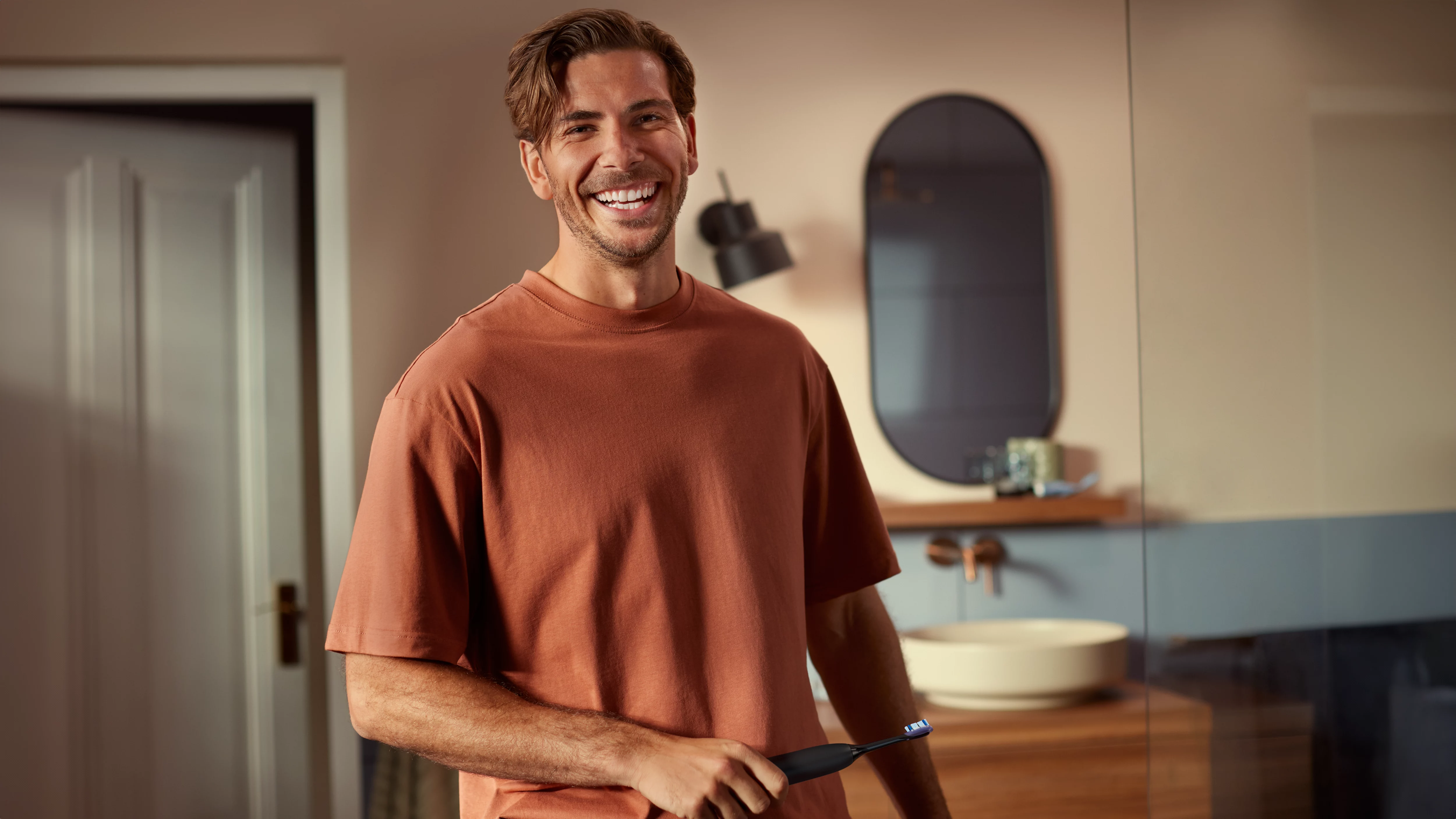 Mann mit Elektrischer Zahnbürste im Badezimmer, lächelnd.