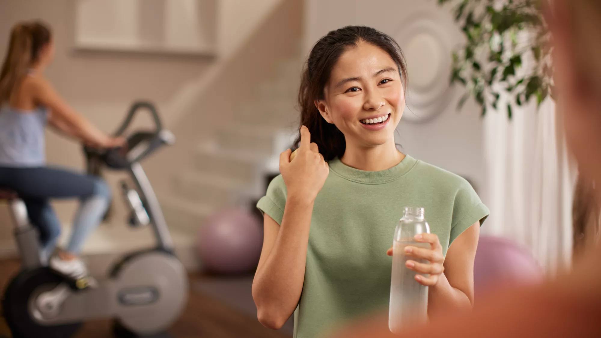 Frau im grünen Shirt hält Flasche. Frau trainiert im Hintergrund auf einem Ergometer.