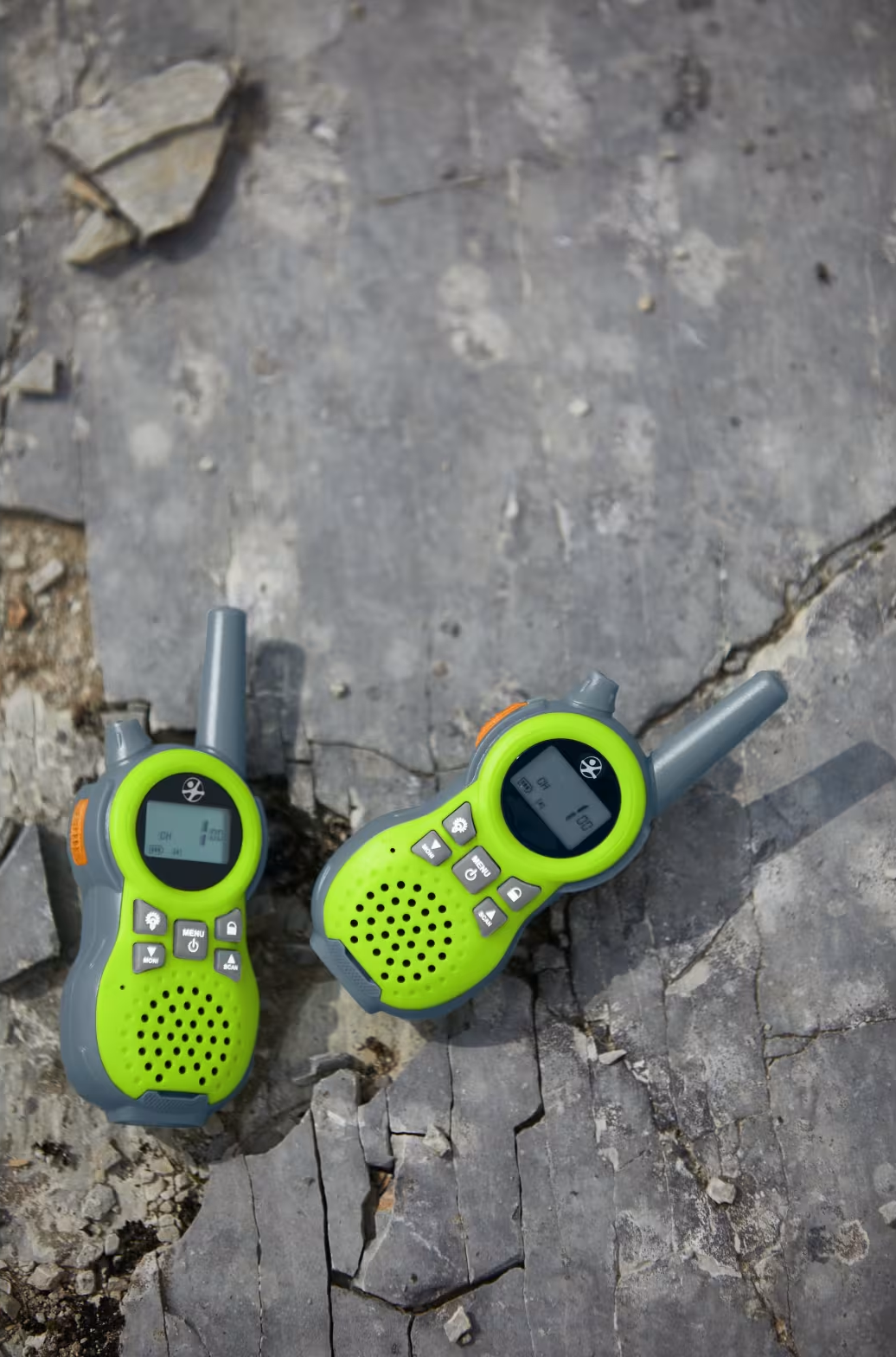 Két zöld és szürke walkie-talkie fekszik egy sziklás, szürke felületen. Az egyik kissé balra, a másik jobbra van.