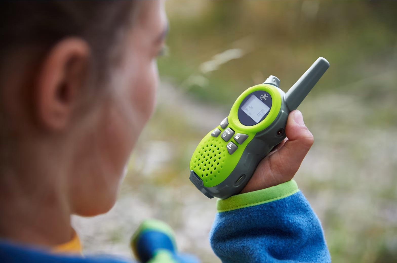 Egy gyerek egy zöld és szürke walkie-talkie-t tart a szabadban.
