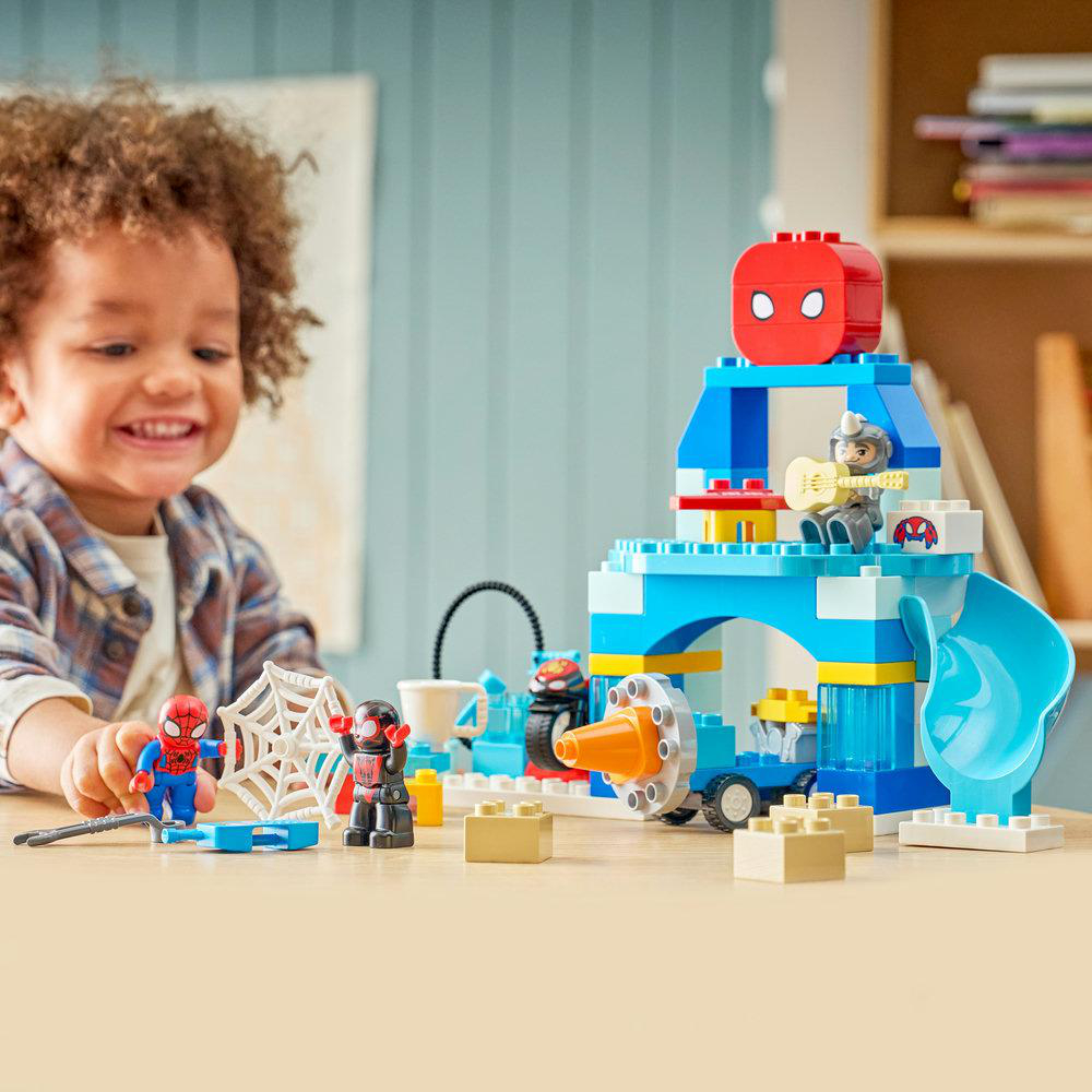 Ein lächelndes Kind spielt mit einem Spiderman LEGO Set mit einer blauen Burg, Figuren und einer Rutsche.
