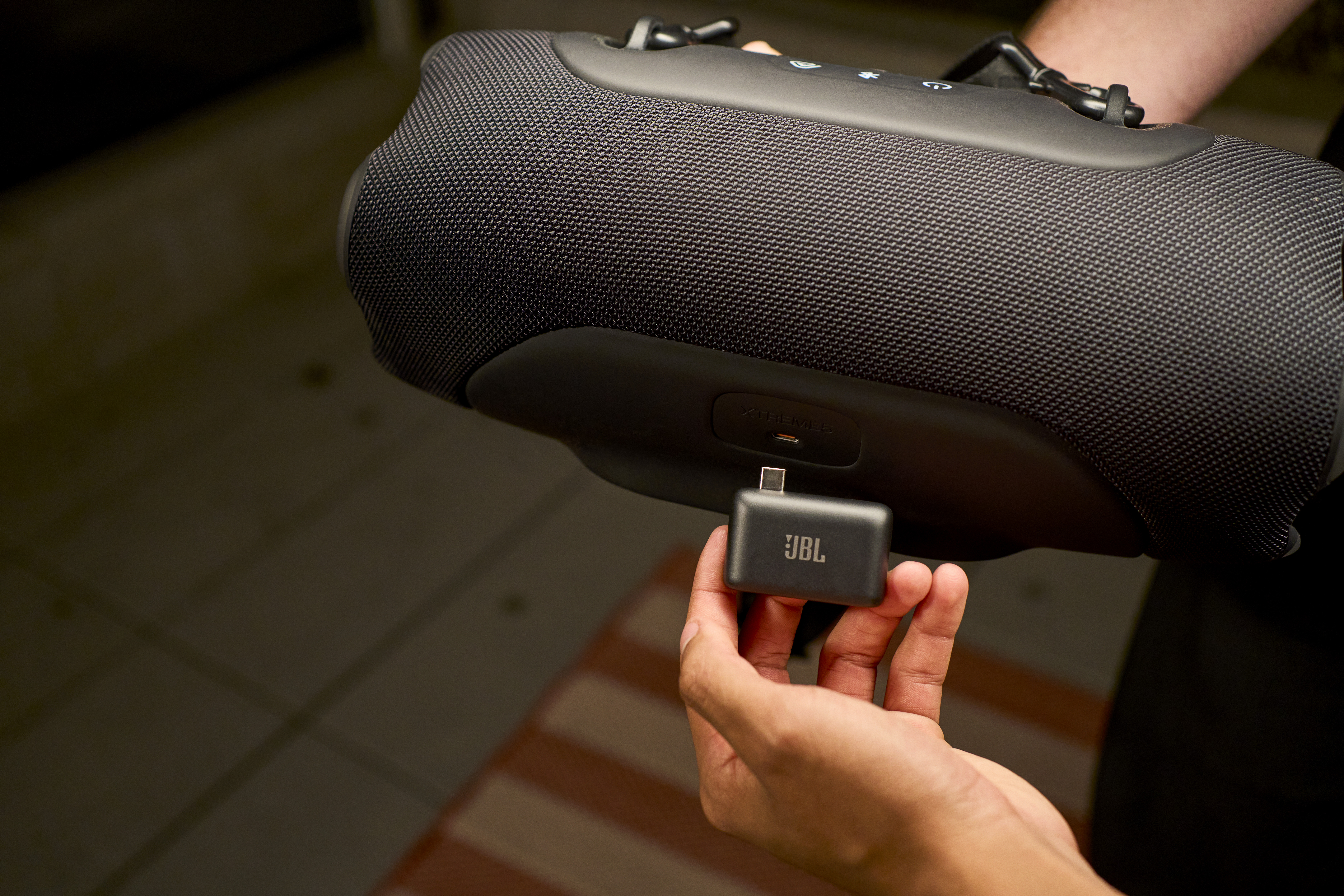 Eine Hand hält einen schwarzen JBL-USB-C-Adapter vor einem schwarzen Lautsprecher.