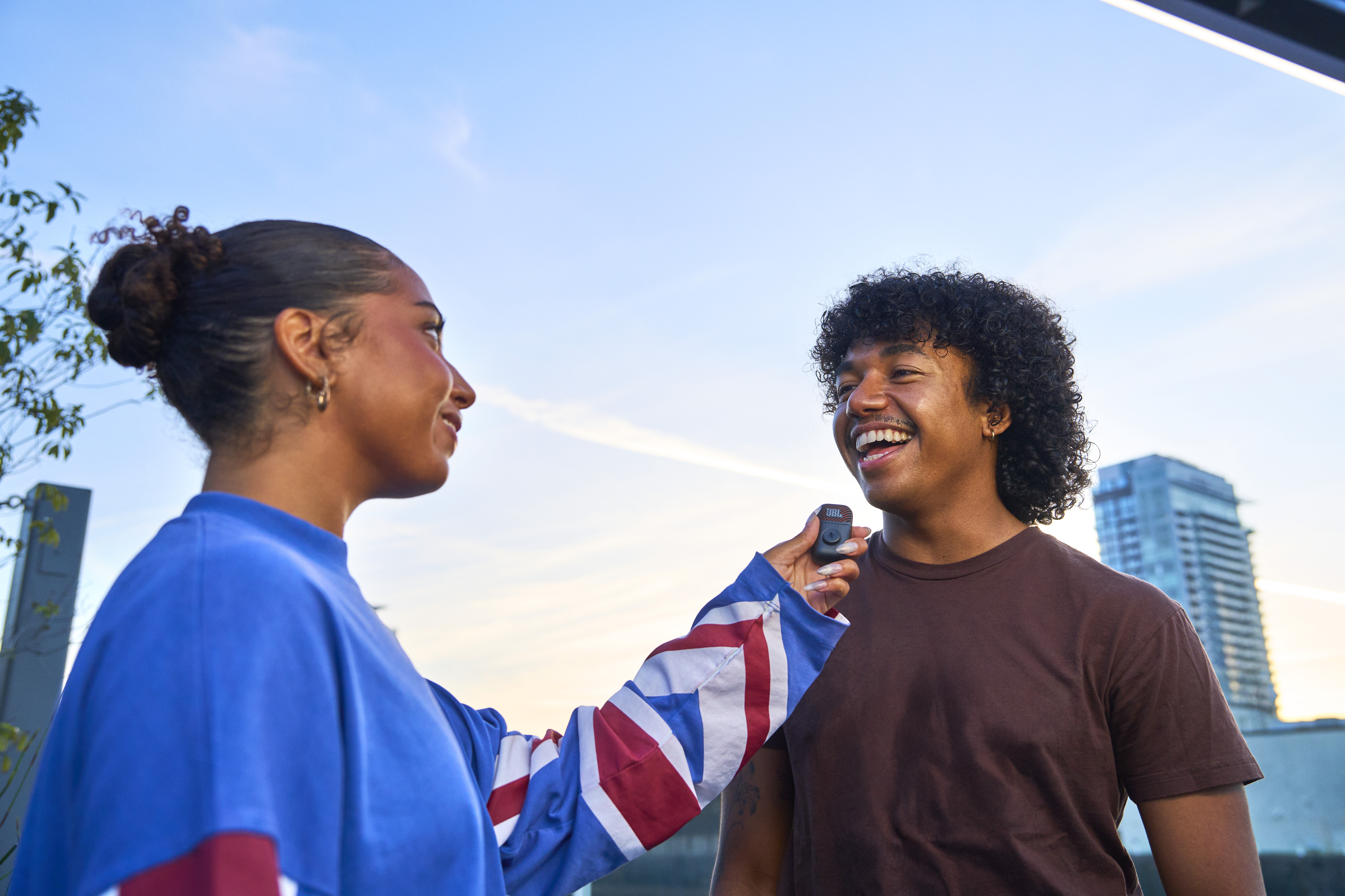 Mann mit lockigem Haar lacht, Frau mit Union-Jack-Ärmel hält ein Gerät.