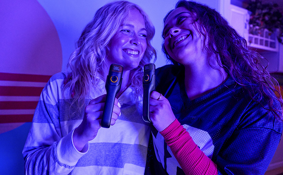 Zwei lächelnde Frauen halten Mikrofone vor blauem und lila Licht.
